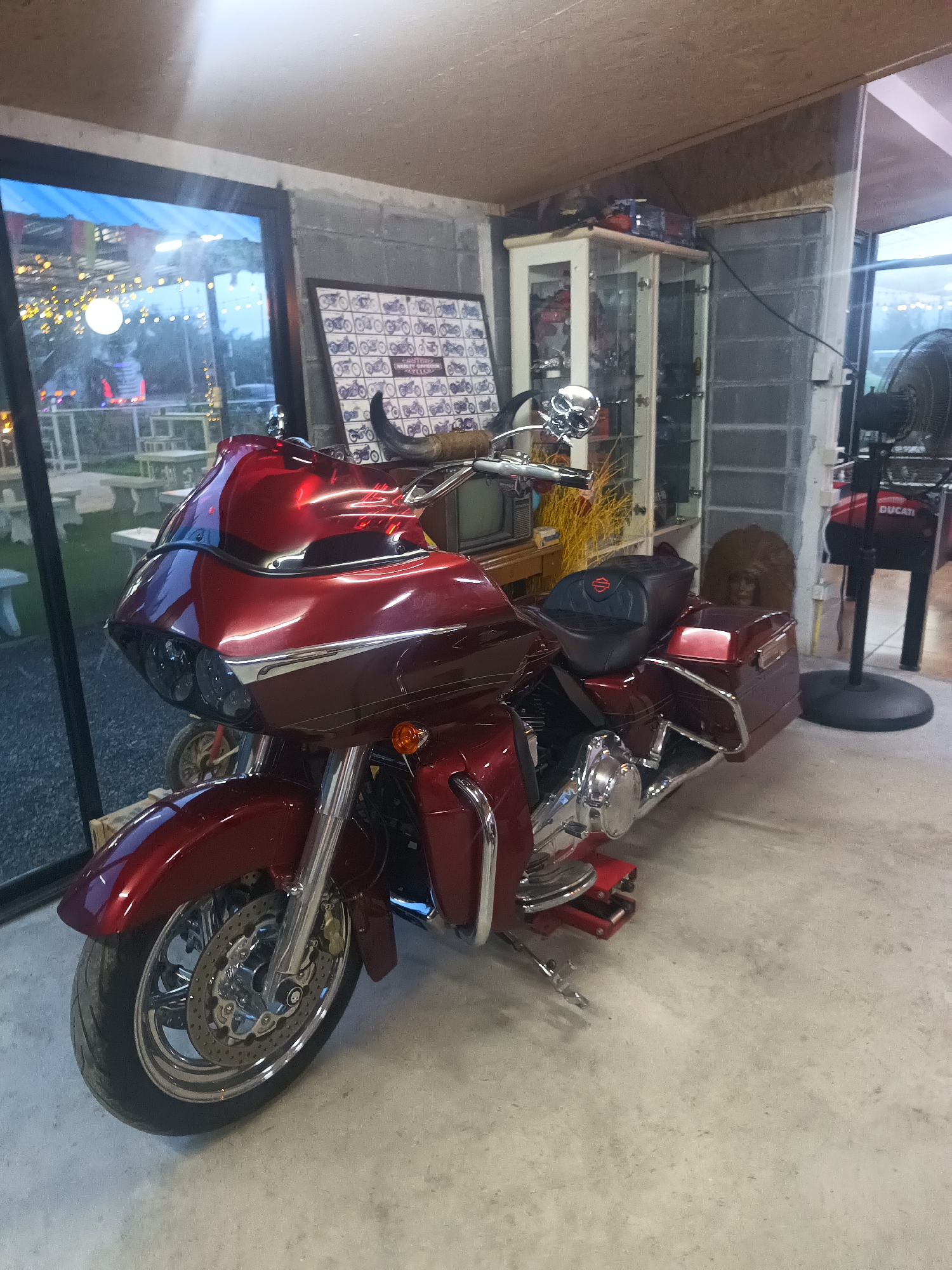 The image depicts a red Harley-Davidson motorcycle parked indoors. Behind the motorcycle, a window reveals an outdoor seating area with tables and chairs. Inside, the space is cluttered with various items, including a glass display cabinet, a television, a poster featuring motorcycle designs, and a Ducati-branded fan. The environment suggests a motorcycle enthusiast's collection or a themed establishment in Surat Thani City Municipality, Thailand.
