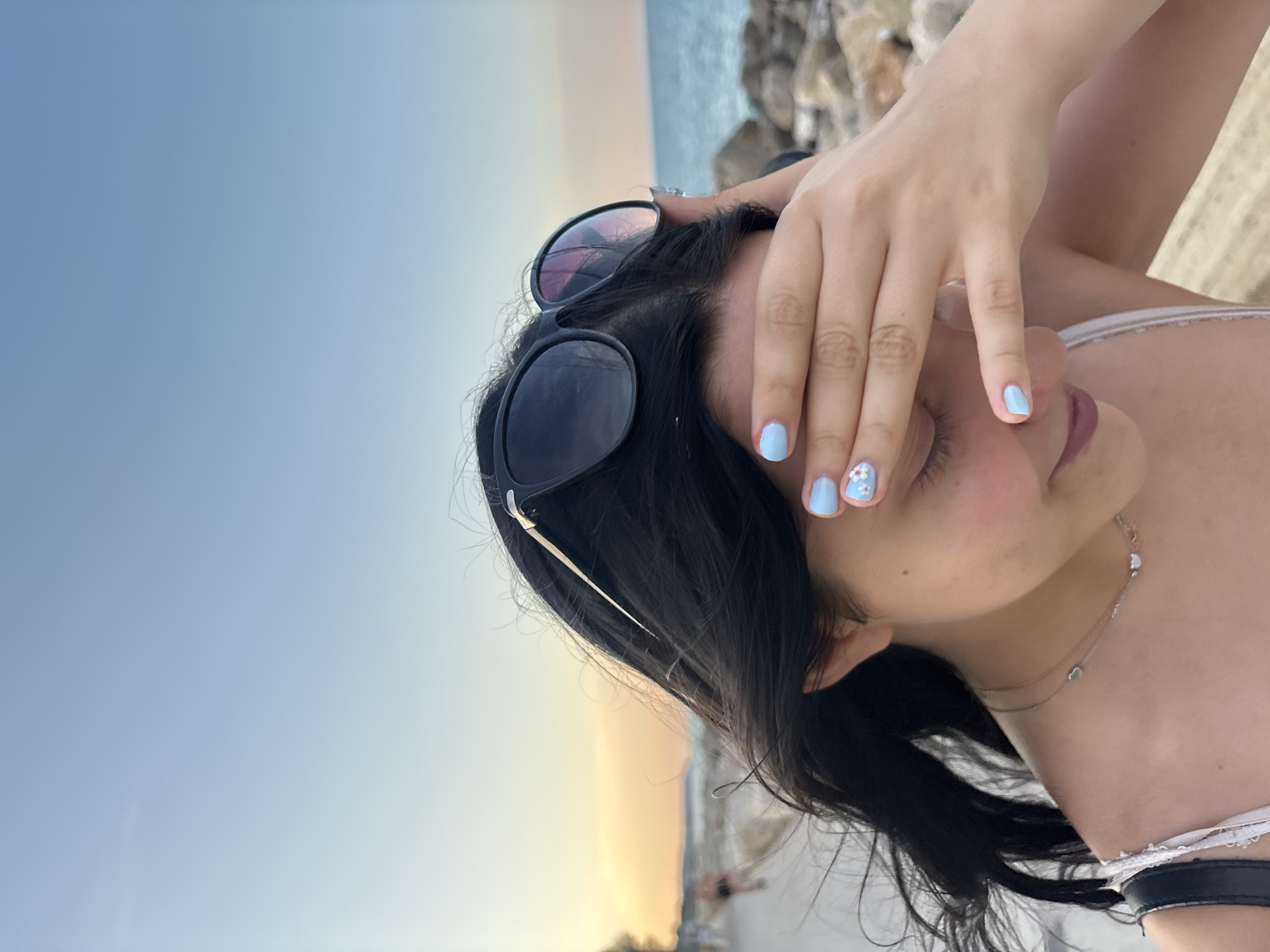 The image depicts a young person covering their eyes with their hand, with sunglasses perched on their head. They are outdoors near a body of water, possibly the Adriatic Sea, with a rocky shoreline. The sky is clear and blue, transitioning to a warm orange hue near the horizon, suggesting it is either sunrise or sunset. The person is wearing a necklace with small heart pendants and a white top. Their fingernails are painted light blue with small white flower designs. The background shows a blurred beach scene with some people and vegetation. The overall setting appears to be a coastal area.