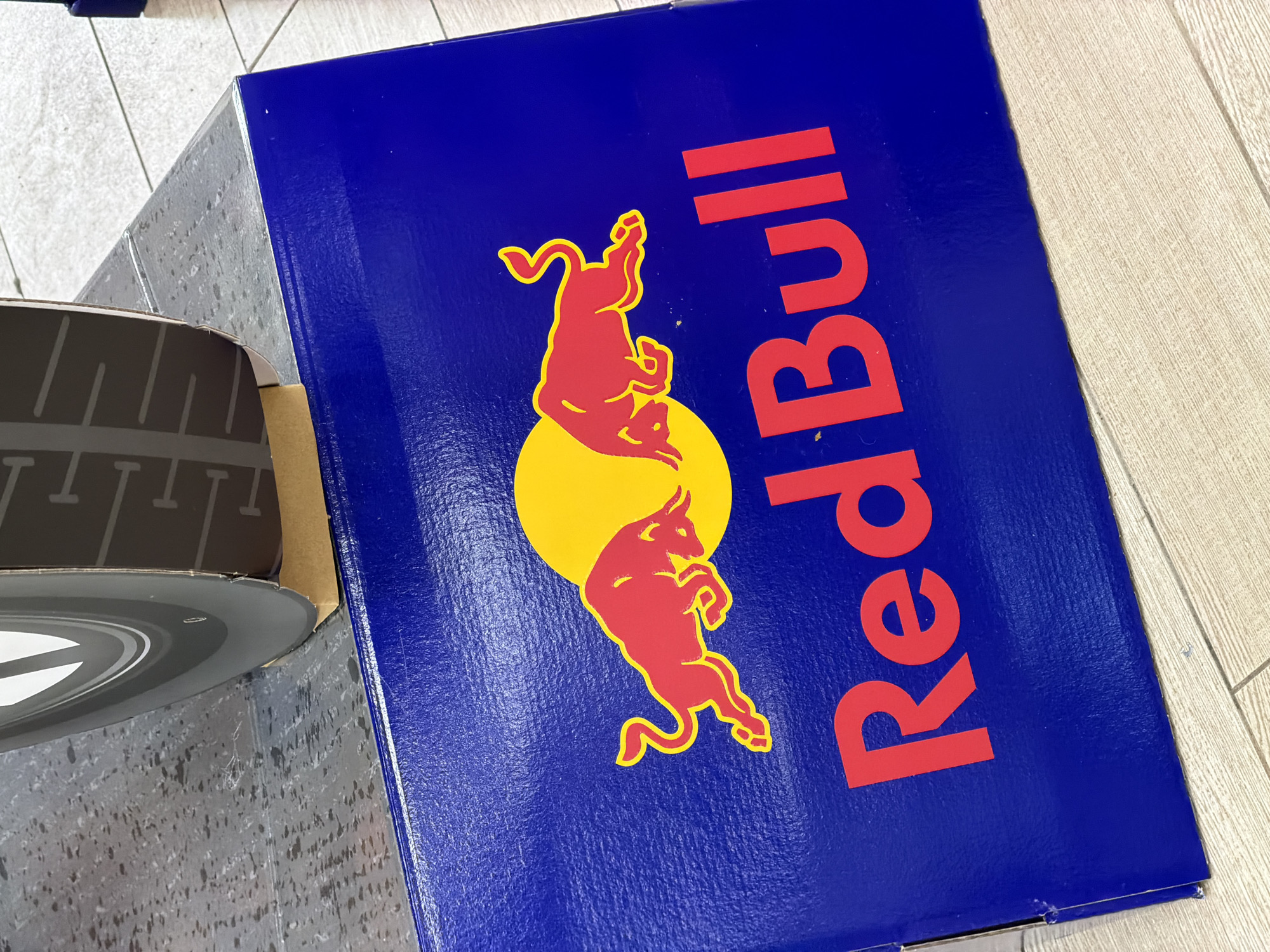 An overhead, close-up, medium shot captures a blue Red Bull branded box. The box is placed on a light-colored wooden floor. On the box, there is the Red Bull logo, which consists of two red bulls charging towards a yellow sun. Below the logo, the words "Red Bull" are printed in large red font. The box appears to be leaning against a large black and grey object which has the texture of a tire. The backdrop consists of a grey, speckled surface. The scene is indoors, with no indication of a specific city or country.