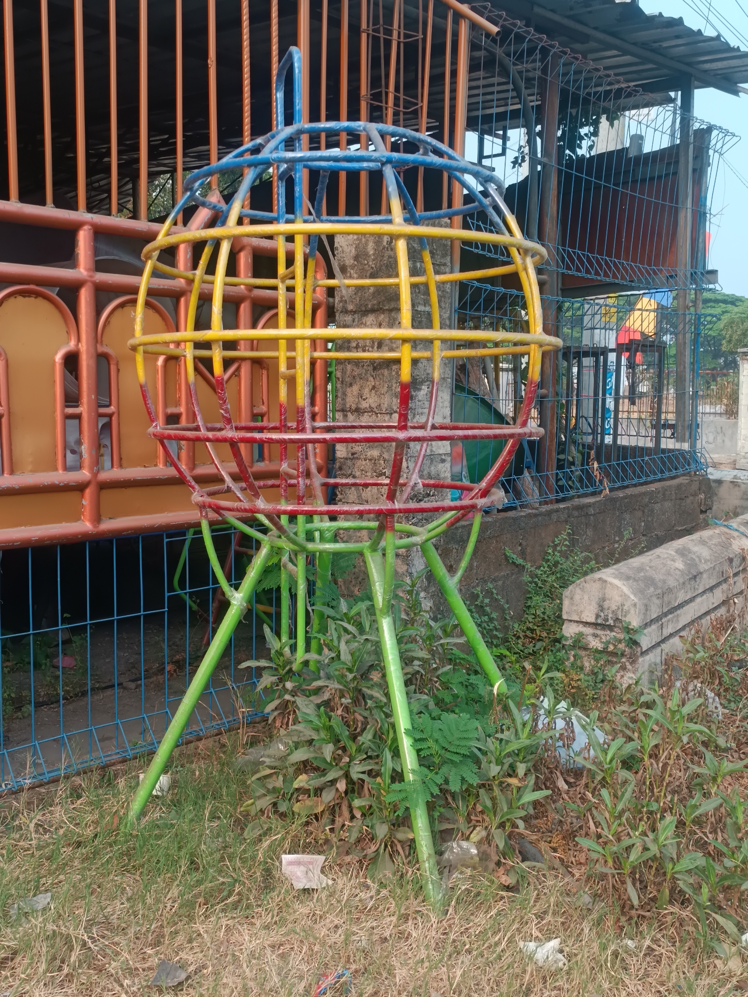 The image depicts an outdoor scene in Sewon, Indonesia. In the foreground, a colorful, spherical jungle gym made of metal bars painted blue, yellow, red, and green stands on grass. The jungle gym has a base with multiple green legs. To the left, an orange metal fence with vertical bars is visible, and behind it, a wall. To the right, a wire mesh fence separates the area from a concrete embankment covered with vegetation and some debris. In the background, there is a roof structure, more fencing, and trees. The ground is covered with dry grass and scattered litter, including paper and plastic. There are no people visible in the image.