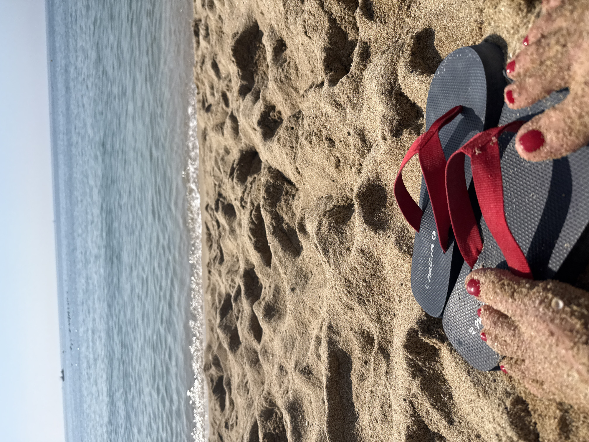 A pair of dark grey flip-flops with red straps are placed on a sandy beach. A person's feet, covered in sand and with red painted toenails, are resting on the flip-flops. The ocean is visible in the background with a clear sky. The location is likely a beach in Barcelona, Spain.