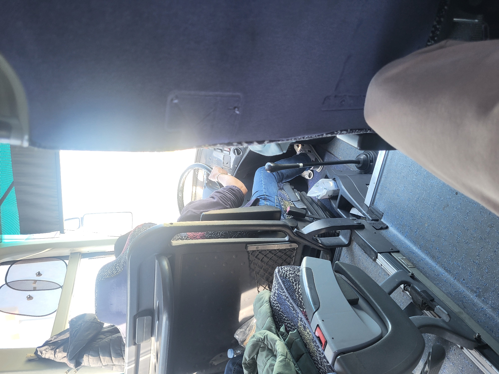 A person wearing jeans and sneakers is driving a bus. The interior of the bus is visible, including the steering wheel, dashboard, seats, and windows. Outside the window, it is bright. Given that this is a bus, the location is likely Orizovo, Bulgaria.