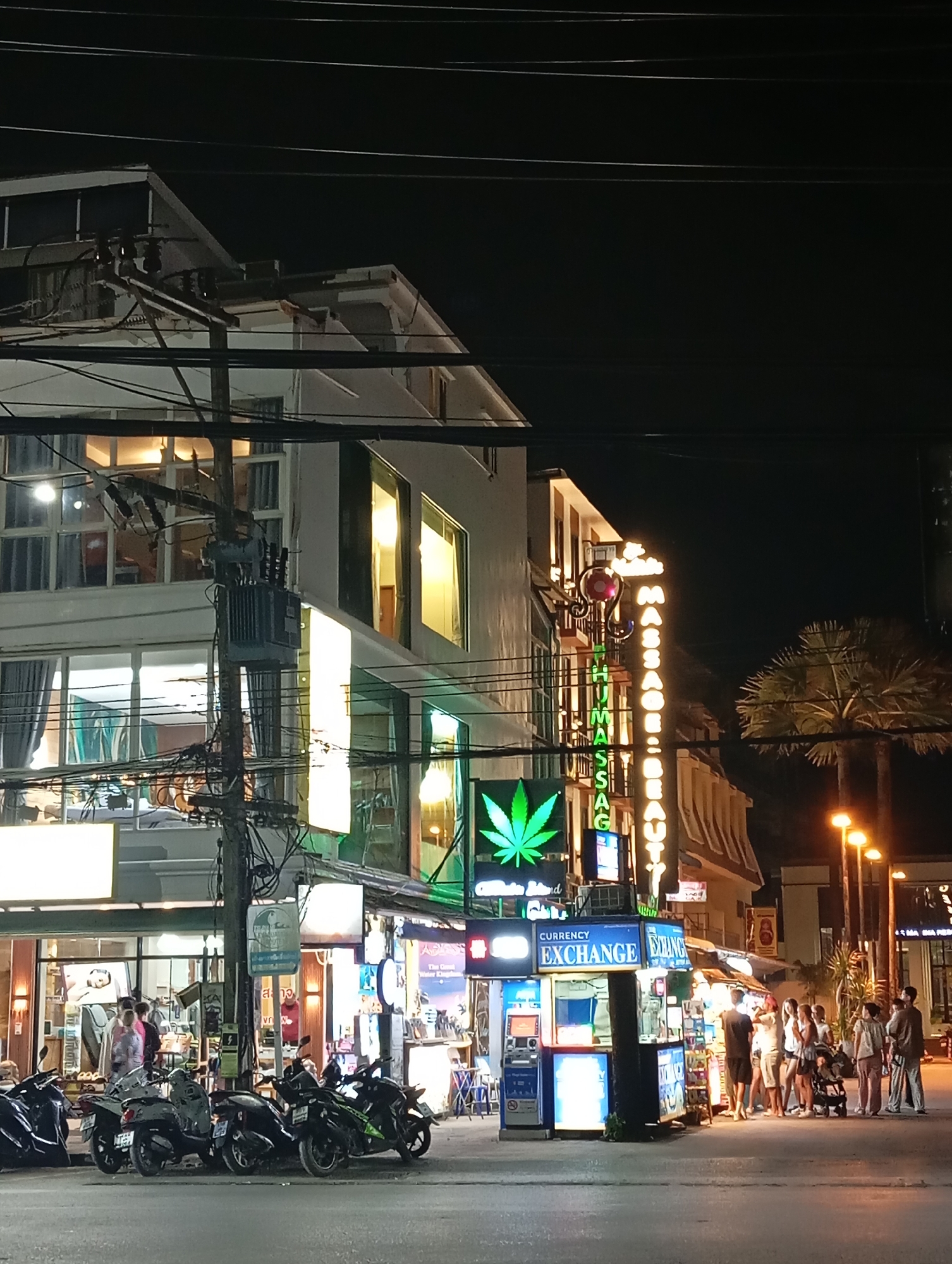 This nighttime photograph captures a street scene in Karon, Thailand, likely a tourist area. The foreground features a row of parked motorcycles. A currency exchange booth is prominent on the right, with several people standing in line or near it. Above the exchange booth, a neon sign reads "MASSAGE & BEAUTY." Further to the left, a building with multiple levels is visible, with illuminated windows and what appears to be a marijuana leaf symbol integrated into a neon sign. Electrical wires and poles crisscross the scene, indicating an urban environment. Palm trees are visible in the background, suggesting a tropical climate. The street is paved, and the overall lighting is artificial, characteristic of a commercial district at night.