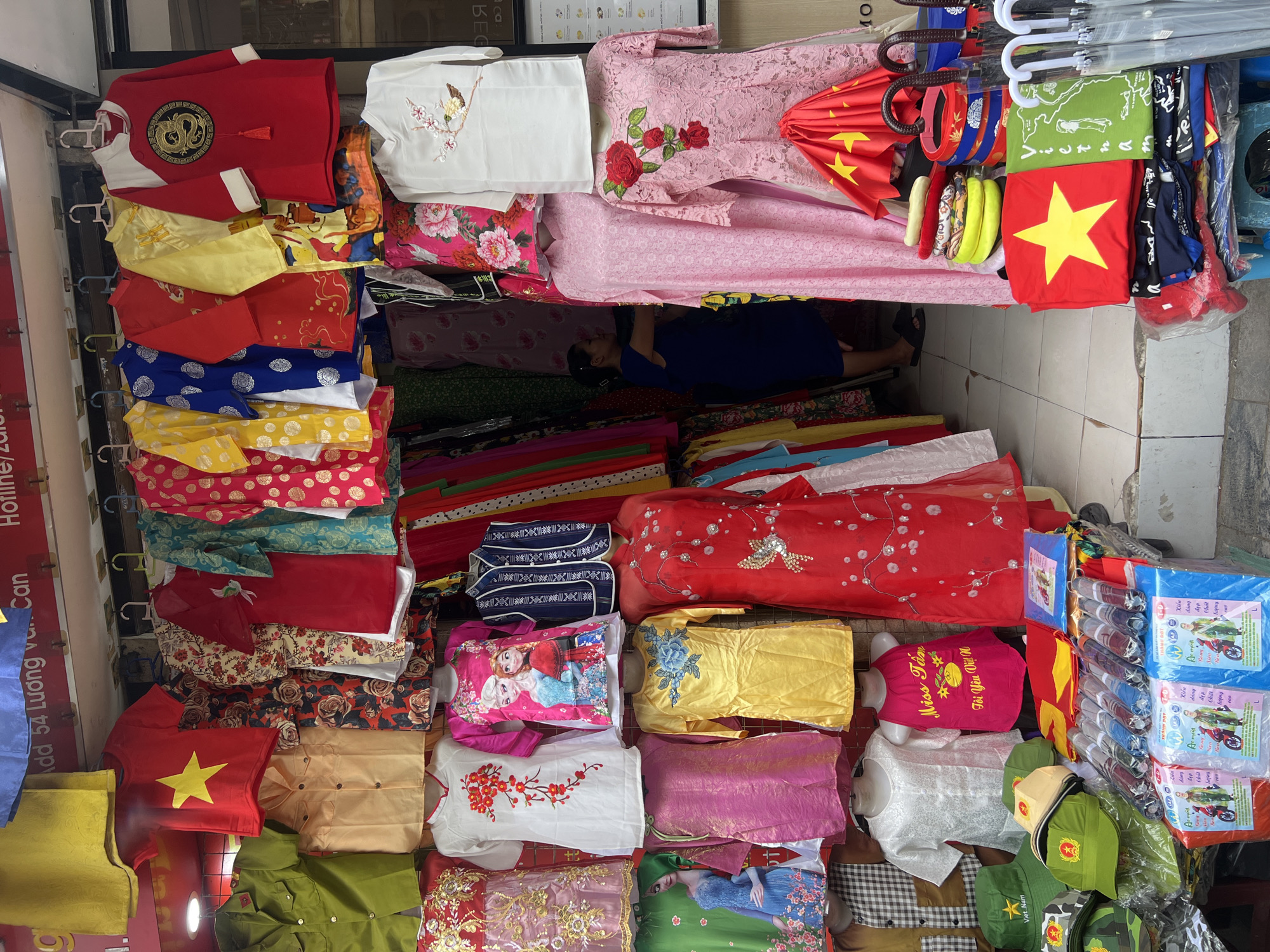 This is a medium shot of a clothing stall in Hanoi, Vietnam. The stall is filled with various types of clothing, including t-shirts, dresses, and hats, many of which are decorated with Vietnamese national symbols. The clothing is displayed on hangers and mannequins, with some items folded on tables. A woman in a dark blue dress is visible in the background, browsing the merchandise. The stall appears to be located on a street, with other stalls and shops visible in the distance. The overall impression is of a busy marketplace, with a focus on traditional Vietnamese clothing and souvenirs.