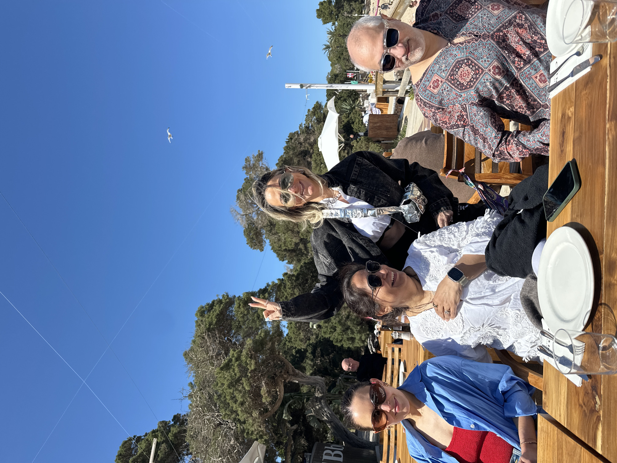 A group of five adults are seated at a wooden table at a restaurant in Sant Josep de sa Talaia, Spain, under a clear blue sky with a few visible wires and birds. From left to right:
A woman wearing a blue shirt over a red top and sunglasses sits at the edge of the table. Beside her, a woman in a white blouse wearing sunglasses gives a peace sign. Next to her, a woman in a white top wearing sunglasses is partially visible. Behind her, a woman in a white top and black jacket wears sunglasses. Across from the women, a man with a bald head and sunglasses wears a patterned shirt.
The table contains plates, glasses, silverware, and a black mobile phone. Trees and vegetation can be seen in the background.