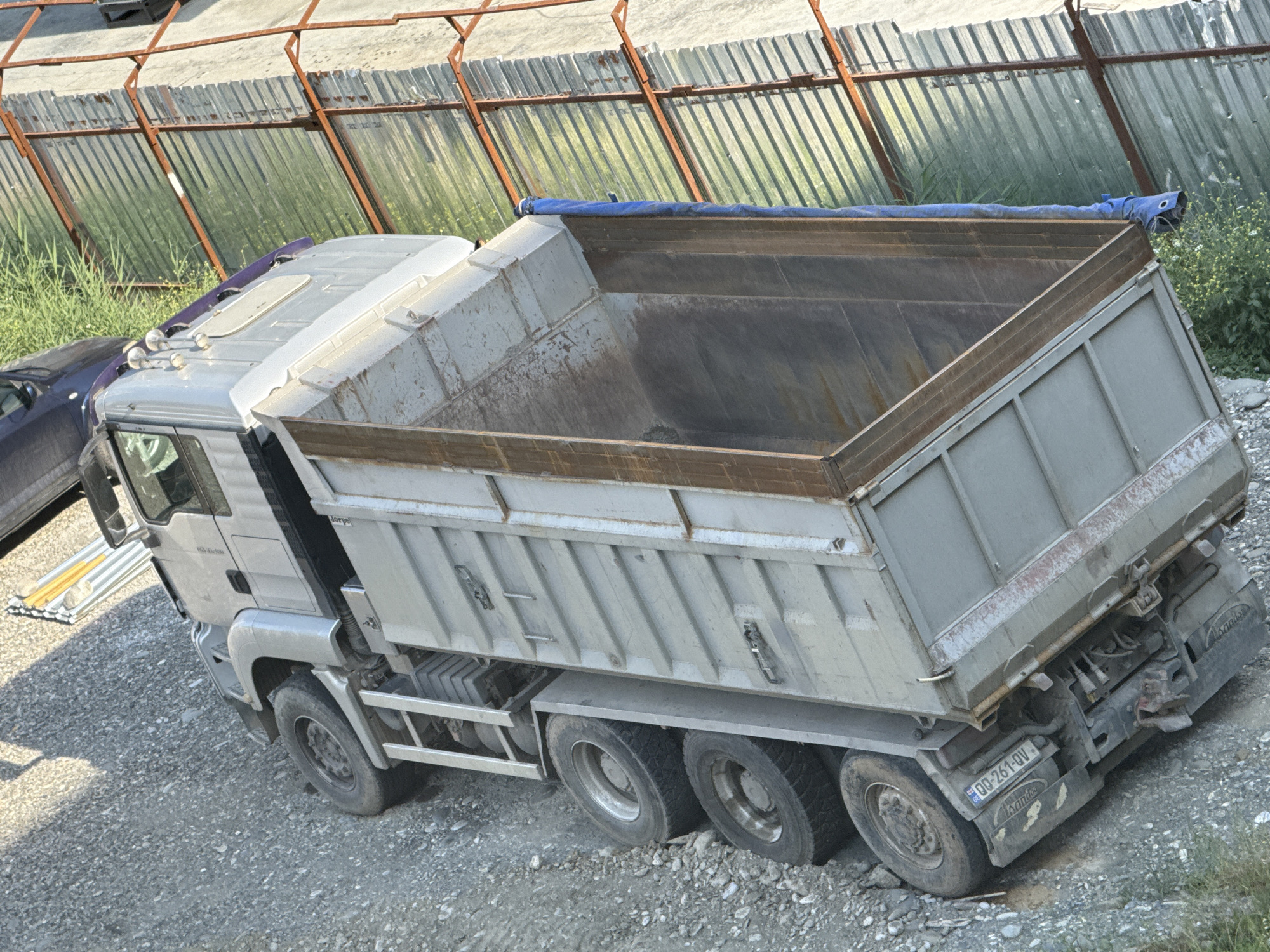 An aerial, high-angle shot captures a dump truck with a raised bed, partially filled with a dark material, parked on a gravel surface. The truck is silver with a weathered, grey dump bed. The bed is covered with a blue tarp. To the left of the dump truck, the front of a dark-colored car is visible. In the background, a corrugated metal fence with orange supports runs horizontally across the frame, backed by a patch of green vegetation. The scene is consistent with a construction or industrial area. The license plate of the dump truck, visible on the rear, reads "QQ-261-QV". The truck appears to be located in the city of Tbilisi, Georgia, suggested by the license plate format.
