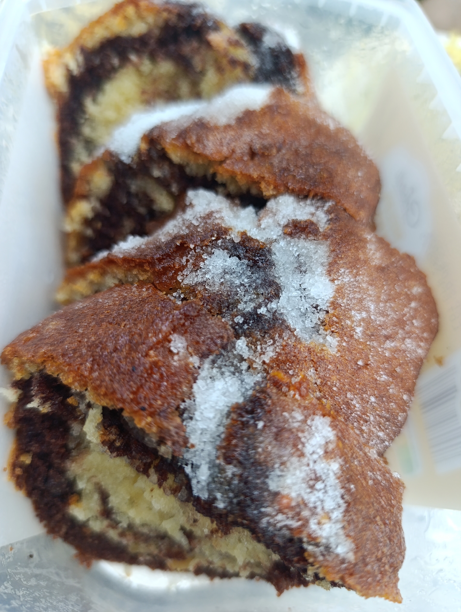 A close-up, high-angle photograph depicts at least three slices of a marble-patterned baked good contained within a translucent white plastic container. The individual slices exhibit an interior composed of light beige or yellow dough intricately swirled with dark brown, likely chocolate or cocoa, dough. The exterior crust of the baked item is a deep, reddish-brown color, displaying a somewhat coarse and irregular texture. Granular white sugar crystals are unevenly distributed over the top surfaces of the slices, creating a prominent sparkling effect. The slices are positioned linearly within the container, which features indistinct grey markings on its right side. The overall lighting is bright, accentuating the texture of the crust and the crystalline structure of the sugar. This scene is located in the city of Nikolichevtsi, Bulgaria.