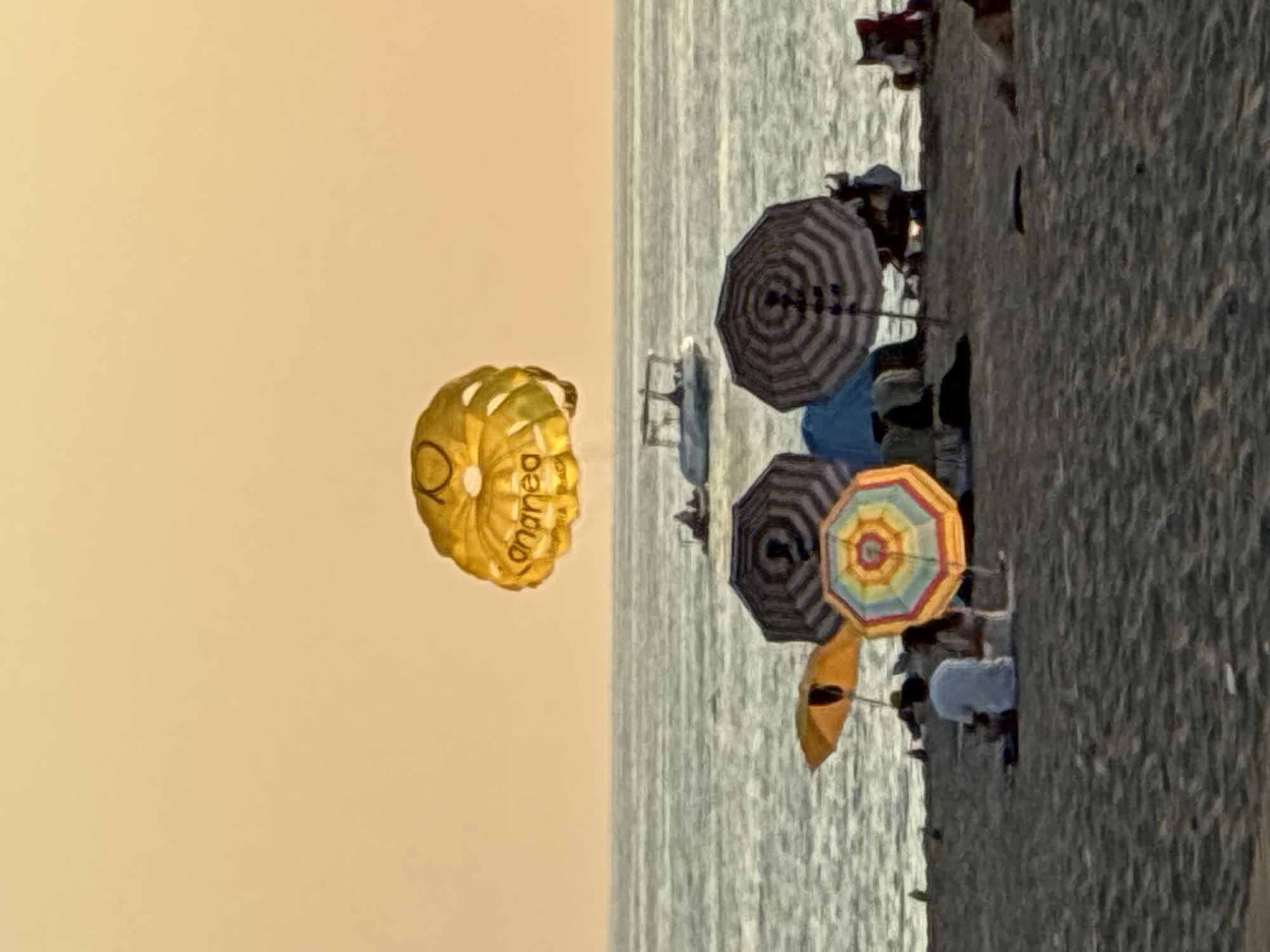 In Alanya, Turkey, a yellow parasail floats in the warm, hazy sky over the Mediterranean Sea. A white speedboat navigates the water below. On the sandy beach, beachgoers are visible, some sitting and relaxing under colorful umbrellas, including a striped umbrella, an orange umbrella, and a multicolored umbrella. The scene is bathed in the soft, golden light of a sunset.