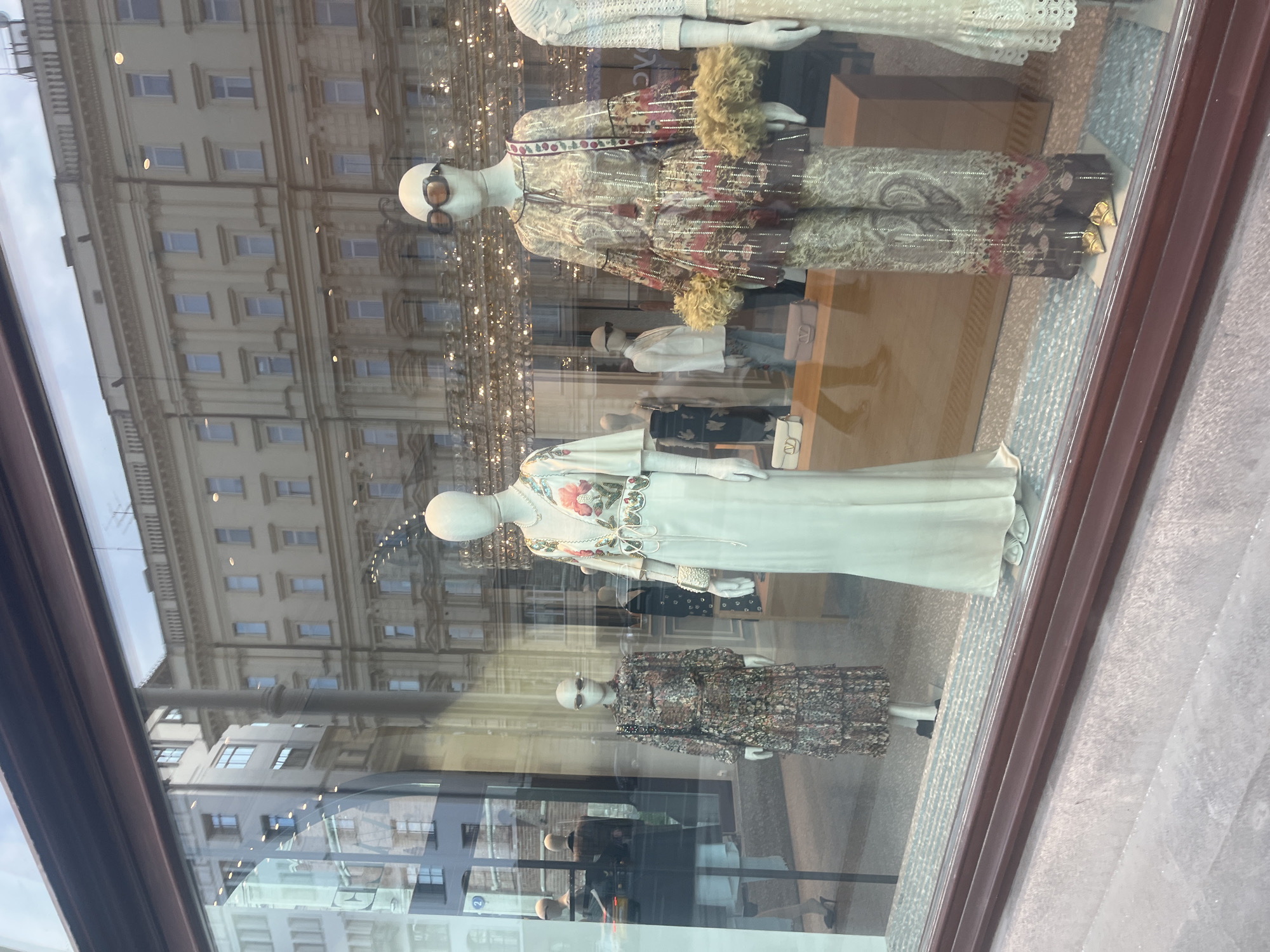 This is a medium shot, eye-level, exterior photograph of a high-end clothing store window display, taken during daylight. The display features multiple mannequins dressed in elaborate designer clothing, showcasing a variety of styles and patterns, including a long, flowing pale green dress with floral embellishments, a patterned long-sleeved dress with ruffles, and a matching printed tunic and pants set with fluffy trim. The mannequins are accessorized with sunglasses and jewelry. Reflections of the city street, including buildings, windows, and streetlights, are visible on the glass of the store window. The building across the street appears to be a historical structure with multiple stories and windows. The pavement in front of the store is visible, showing a textured, stone-like surface. The scene suggests a retail environment in a city center, likely a fashion district, given the nature of the displayed merchandise and the urban backdrop. Without additional context, the specific location in Moscow, Russia, cannot be definitively identified from the image alone.