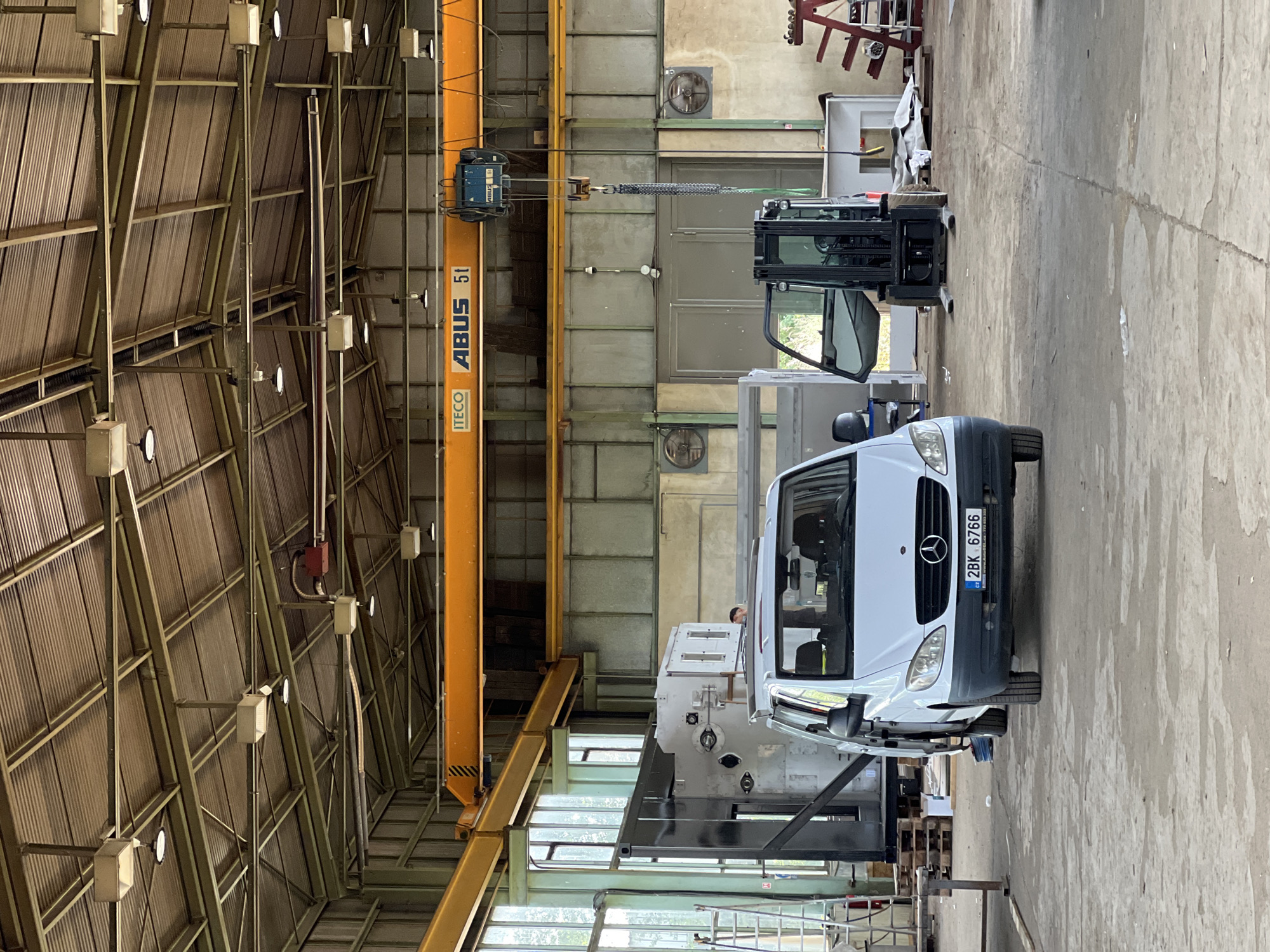 This is an interior, eye-level, medium shot of a large industrial workshop or warehouse in Lanžhot, Czechia. The space is filled with machinery and equipment. A white Mercedes-Benz Vito van is parked in the foreground, facing towards the viewer, with its license plate reading "2BK 6766". To the right of the van, a black electric forklift is positioned with its driver's side door open. In the background, a large overhead crane with the branding "ITECO ABUS 5t" is visible, suspended from the ceiling. Various industrial components, including large metal structures and pallets, are scattered throughout the space. The ceiling is constructed with corrugated metal roofing supported by exposed steel beams. Natural light streams in from large windows on the left side of the image. One person is partially visible behind a large piece of machinery in the mid-ground. The overall environment appears to be a functional, operational industrial setting.