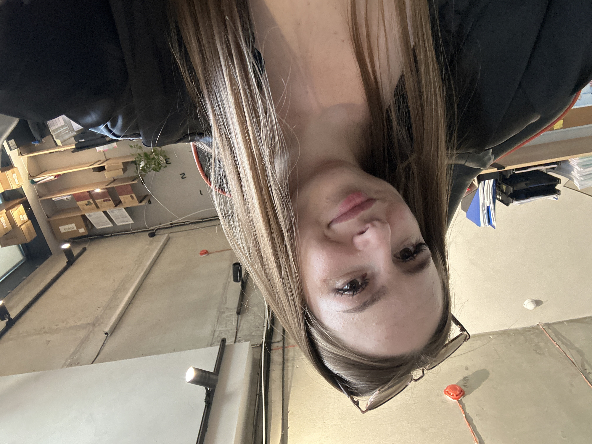 A selfie of a young woman with long brown hair and glasses is taken from a low angle. She is wearing a black jacket. The background appears to be an office or workshop with exposed concrete ceilings and walls, industrial lighting fixtures, and shelves filled with boxes and office supplies.