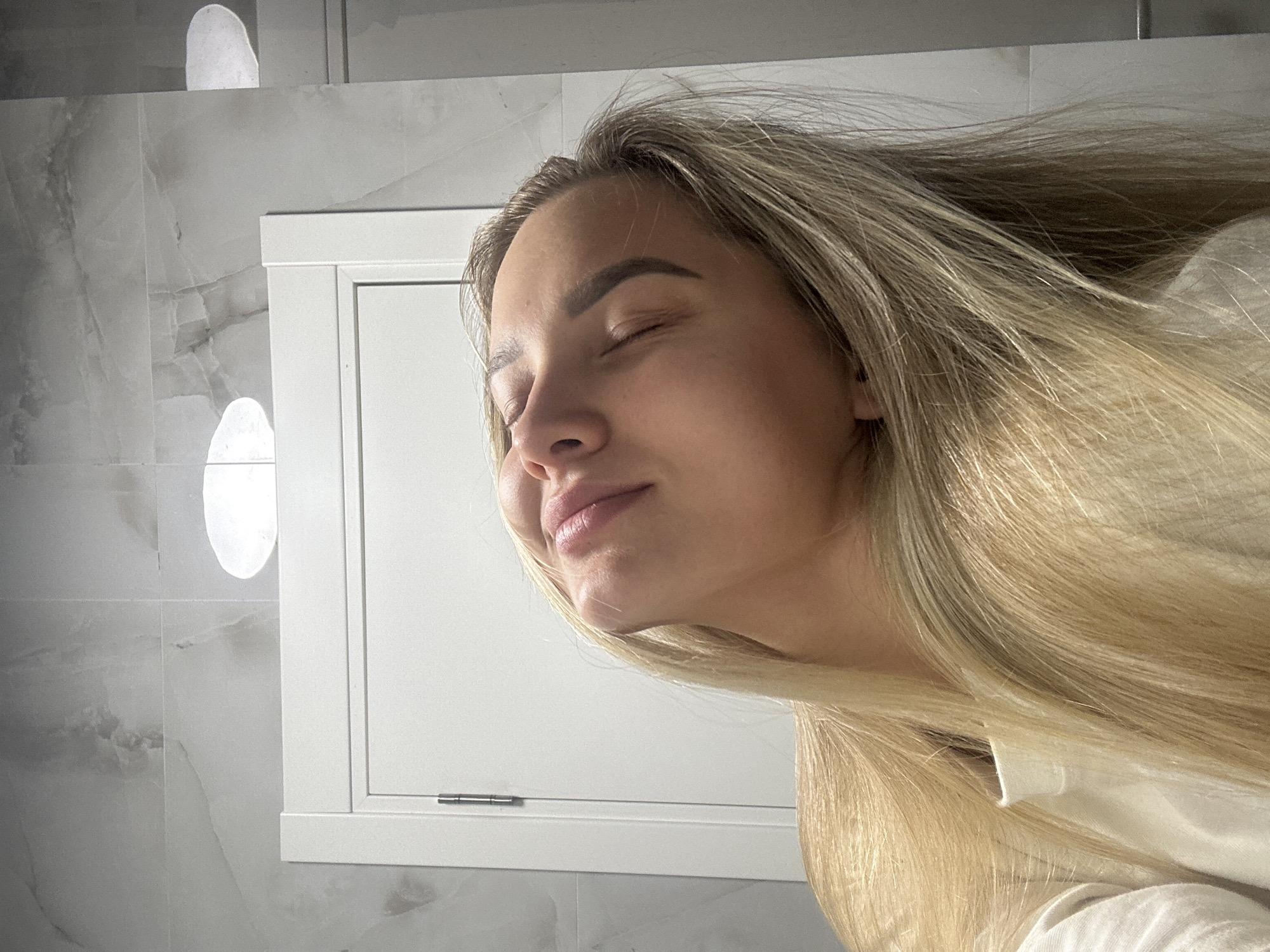 A young woman with fair hair and closed eyes is depicted in a close-up shot. She appears to be indoors, possibly in a bathroom, given the tiled wall and cabinet behind her. The location in Budva, Montenegro is not evident from the image.