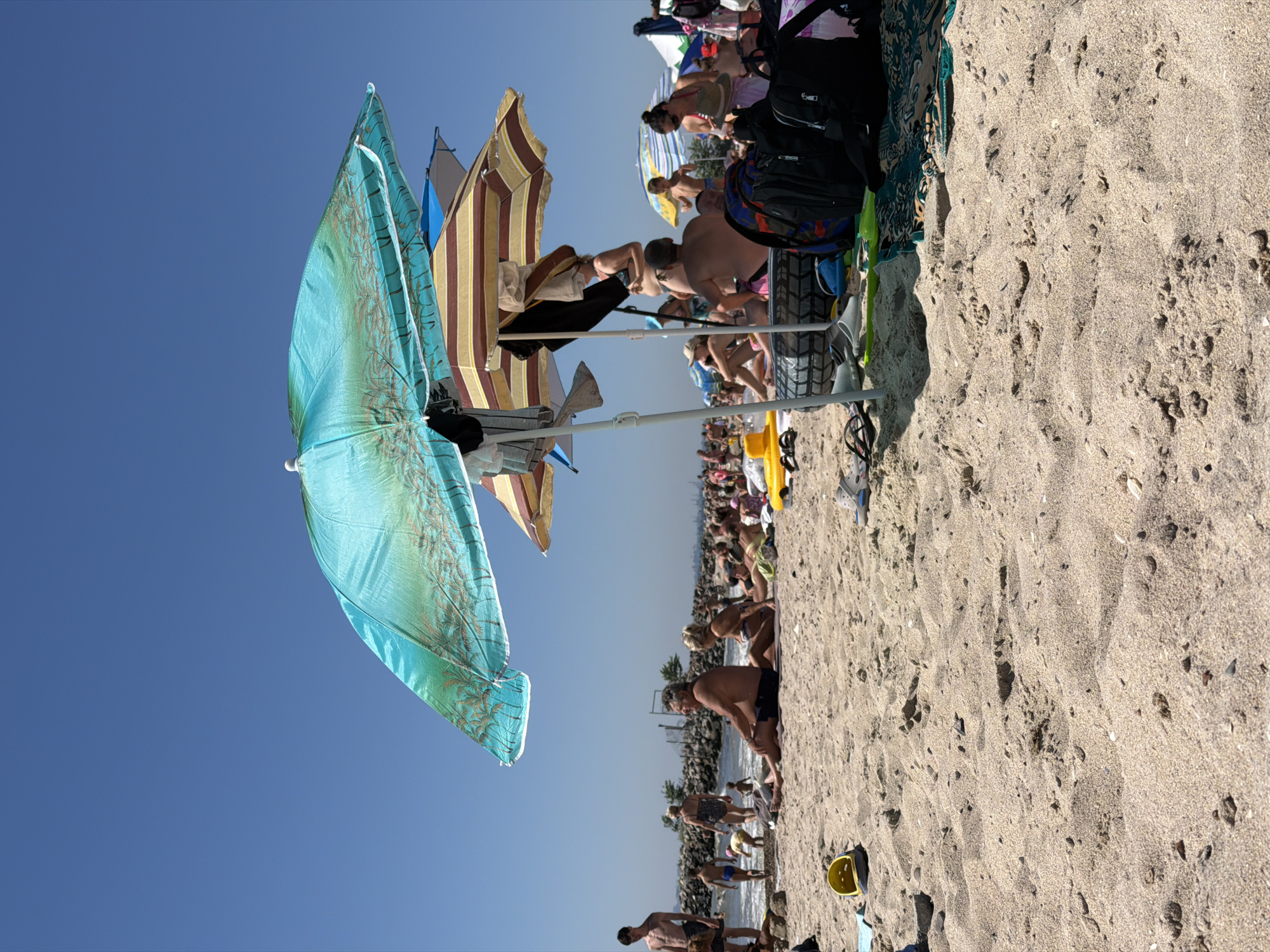 This is a photograph taken on a sunny day at a crowded beach in Odesa, Ukraine. The foreground is dominated by sandy beach with several pairs of flip-flops and a yellow child's bucket. In the mid-ground, people are seated and lying on the sand, some under beach umbrellas. A large turquoise umbrella with a palm tree pattern is prominently featured in the upper part of the frame, layered over a striped umbrella. Several backpacks and beach bags are visible near the umbrellas. In the background, more people are seen walking and wading in the shallow water. A breakwater structure made of large stones extends into the sea. The sky is clear and blue.