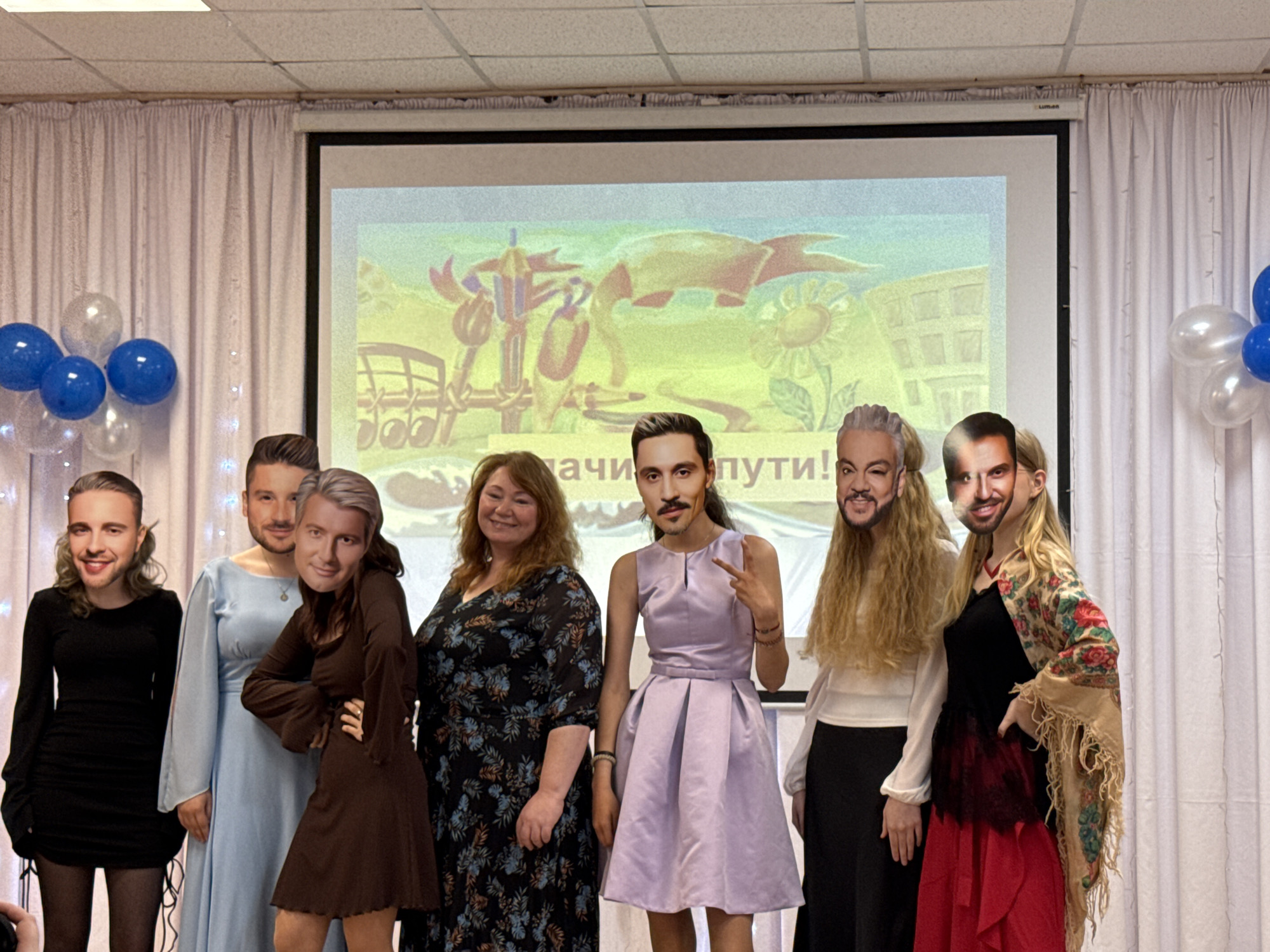 This indoor group photograph, taken in Foky, Russia, features eight individuals, predominantly women, posing for the camera. Each person is holding a cutout mask depicting a male face in front of their own, adding a humorous and playful element to the scene. The attire of the participants varies, including a black long-sleeved dress, a light blue long gown worn by a person with a masculine mask, a brown short dress, a dark floral pattern dress, a lavender satin dress, a white top paired with a black skirt, and a black top with a red tiered skirt and a patterned shawl.
The setting appears to be an event hall or banquet room, characterized by white floor-to-ceiling curtains, standard ceiling tiles, and decorations such as clusters of blue and clear balloons flanking a large projector screen. The screen displays a colorful, stylized image featuring elements like a sun, buildings, and musical notes, along with partially visible Cyrillic text that reads "...ачи пути!", suggesting a phrase like "Good luck on your path!". The artificial lighting indicates an indoor evening or daytime event. The use of masks and the peace sign gesture by one individual convey a celebratory, informal, and fun atmosphere, possibly for a party, performance, or a special celebratory gathering.