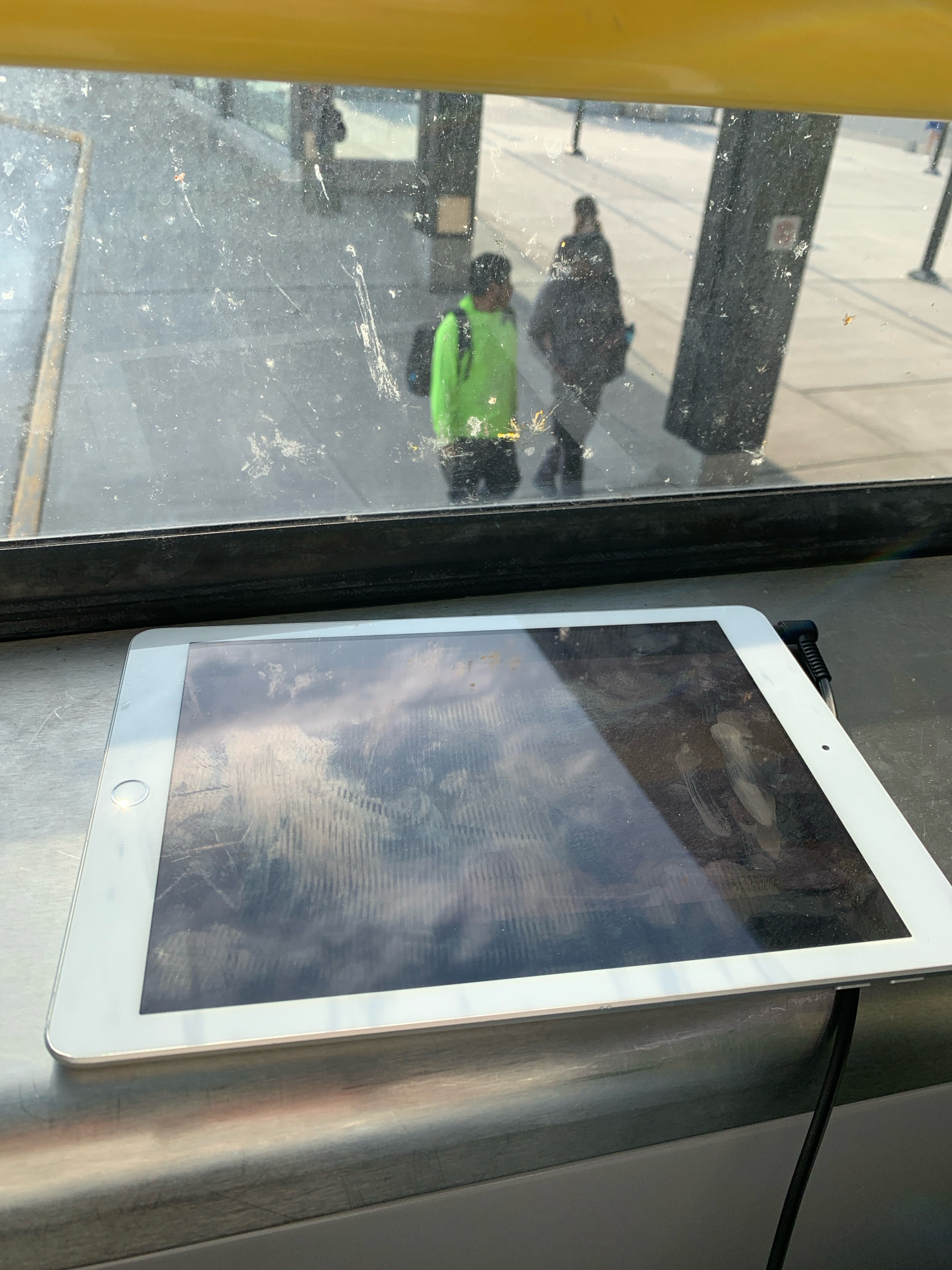 The image shows an iPad on a metallic surface, possibly a counter or table, with a charging cable plugged into its right side. Reflected on the iPad's screen are clouds and a ridged structure. Behind the iPad, a dirty glass window overlooks an outdoor scene. Through the window, two individuals are visible. One person, wearing a bright green jacket, stands to the left, while another person, wearing dark clothing, is to the right and further back. The environment appears to be a public transit waiting area or a similar urban setting, possibly at a bus stop or a train station. The overall impression is of an indoor space with a view of the outside.