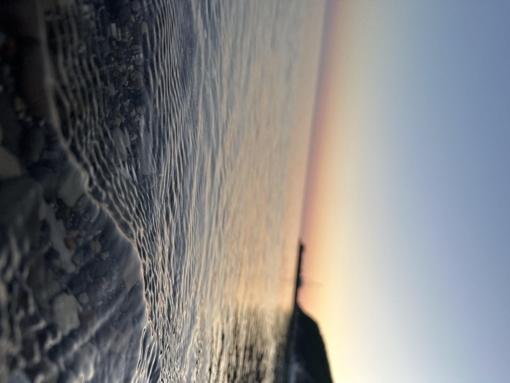 The image shows a shoreline at sunset in Novomikhaylovsky, Russia. The foreground is a close-up view of clear water rippling over pebbles. The background shows the sun setting over the water, and the sky is a gradient of colors from orange and yellow near the horizon to blue. In the midground, there's a dark, blurry object.