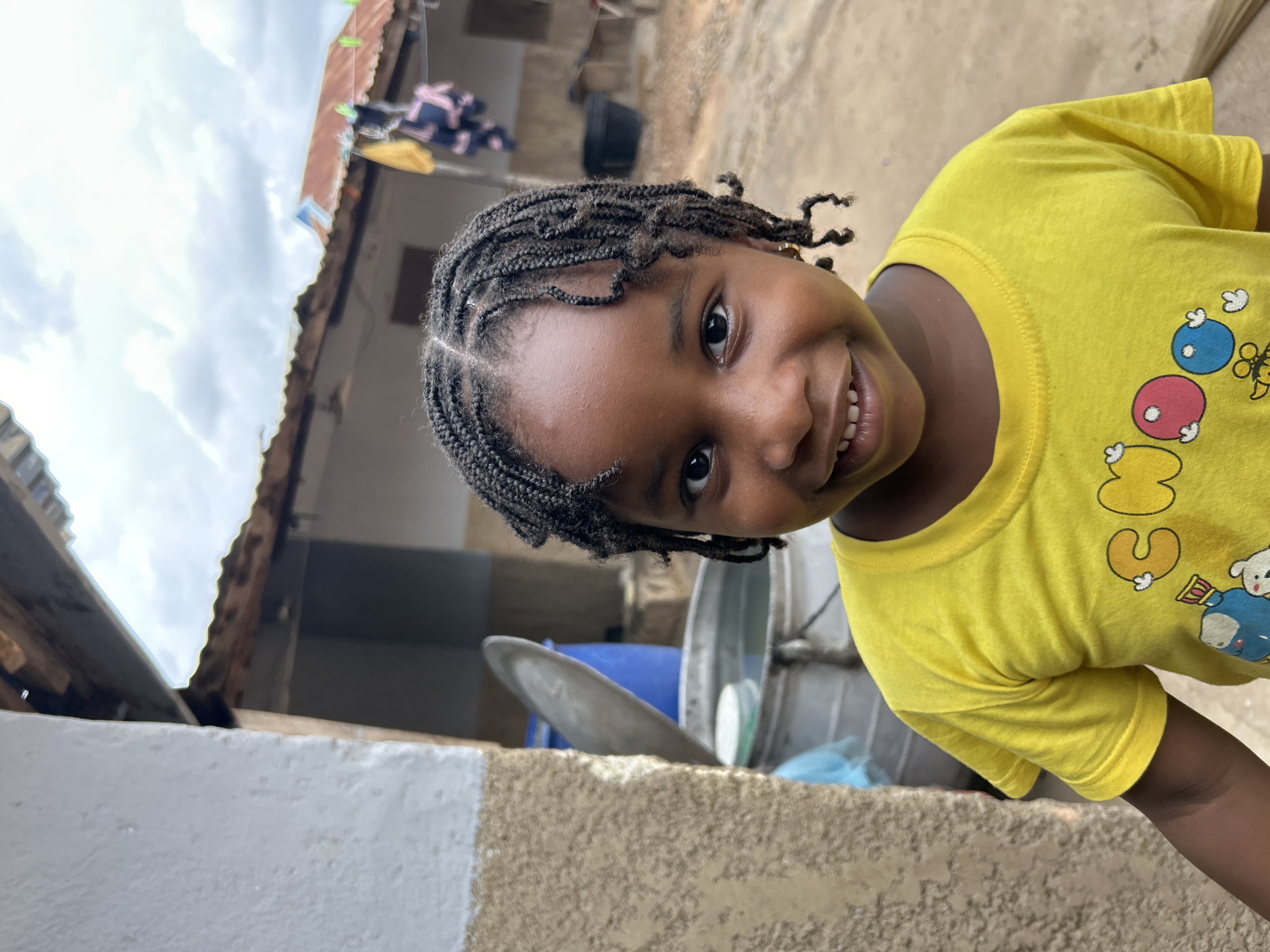A young girl with braided hair and a yellow t-shirt stands smiling in an outdoor environment. In the background, there's a building with a visible roof, clothing hanging on a line, and large metal containers. The foreground consists of a wall with a two-tone paint job. The photo was most likely taken in Jos, Nigeria.
