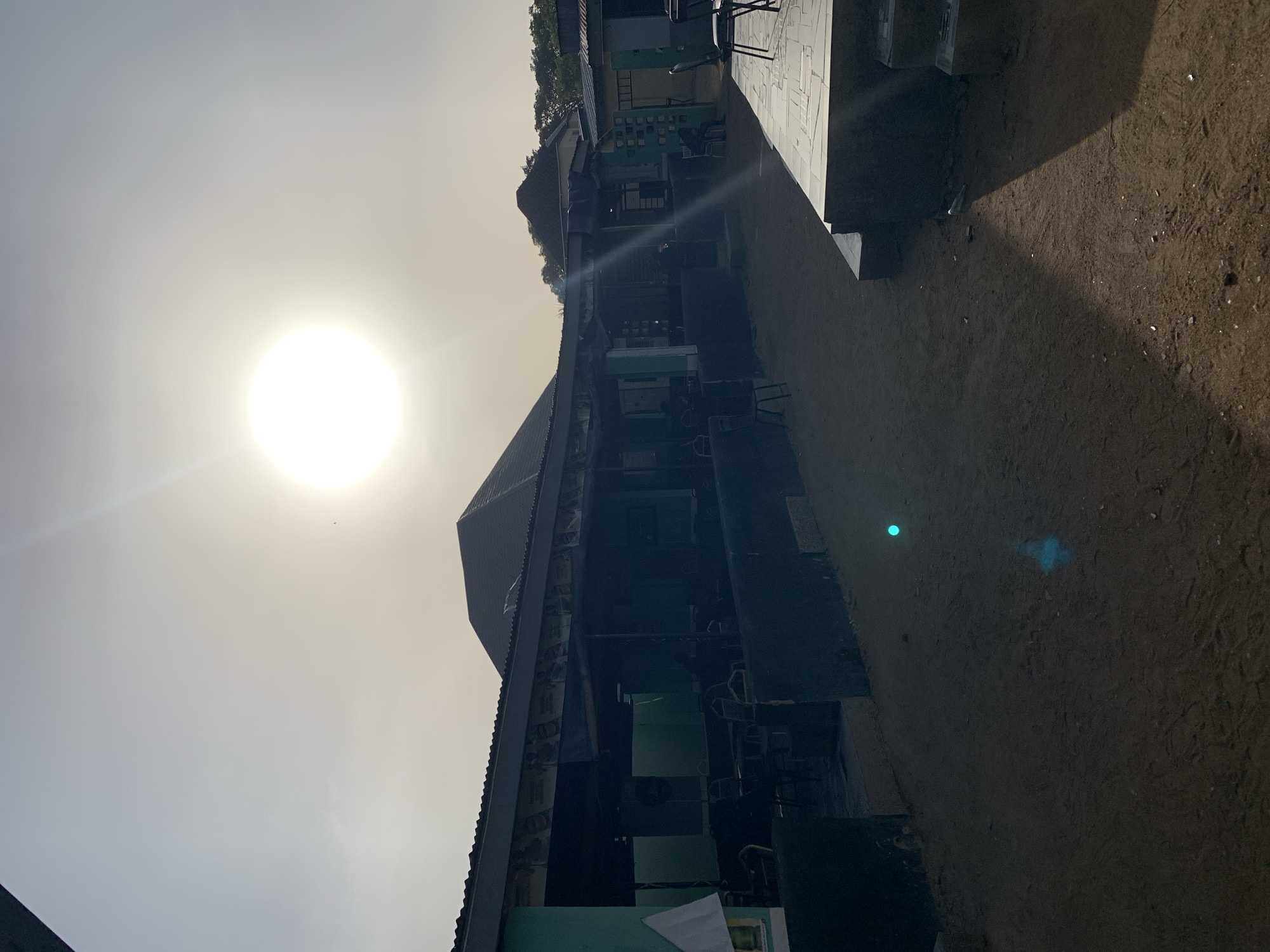 The image shows an exterior view in New Stadium, Nigeria, on a bright, sunny day. The sun is high in the sky, causing significant glare and obscuring some details. The architecture appears to be a row of commercial buildings. The buildings are constructed from metal and cement. The surrounding ground is a mix of dirt and sand, littered with debris. The scene appears to be a street or open area adjacent to the buildings, and there are chairs visible inside of the commercial buildings.
