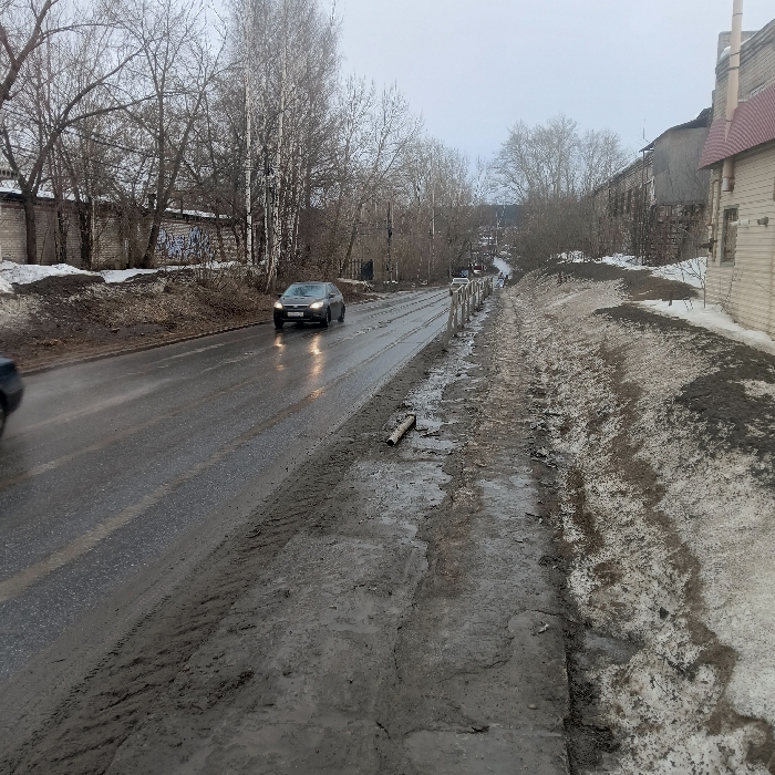 The image displays a wet, two-lane asphalt road in Chaikovsky, Russia, captured during late winter or early spring under an overcast sky. The road exhibits signs of thaw, with accumulated water and visible tire tracks, and descends into the distance, curving slightly to the right. A dark-colored sedan, with its headlights illuminated, is traveling towards the viewer in the left lane; its front license plate, M163KA 159, is partially legible. A second, dark-colored vehicle is partially visible on the far left edge of the frame, moving away from the viewer.
To the immediate right of the road, an unpaved shoulder consists of mud, melting snow, and ice. A light-colored, cylindrical object lies horizontally on this shoulder. A weathered metal guardrail or fence runs parallel to the road along this side. Beyond the shoulder, a dirt embankment covered with patches of snow and mud rises, partially obscuring a light-colored building with a red gabled roof located further right. In the background on the right, additional structures and bare trees are visible.
On the left side of the road, a higher dirt embankment, also mixed with snow and mud, is present. Behind this embankment stands a grey concrete or brick wall covered with graffiti. Numerous bare deciduous trees, possibly birches, are positioned behind the wall and along the left side of the road, with more trees and distant buildings visible further down the road. No individuals are present in the scene.