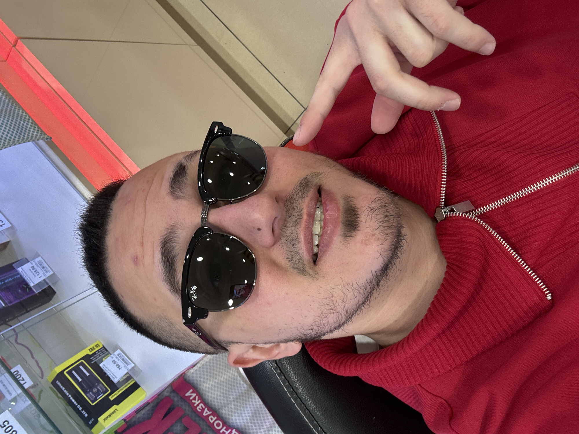 Here's a concise description of the image:
A man with short dark hair, wearing sunglasses and a red zippered sweater, is positioned in the image with his face oriented towards the camera. He is gesturing with his hand. The background includes a display case featuring various items, including packaged electronic components with Cyrillic lettering ("ЦНОРАЗКИ") and pricing ("760,00", "1 125,00"). A brand name, "LiitoKala," is also visible. The sunglasses are identified as "Ray-Ban." Based on the Cyrillic script and the presence of the "LiitoKala Power CO. LTD," and "ЦНОРАЗКИ" on the packaging in the background, the location is Saint Petersburg, Russia.