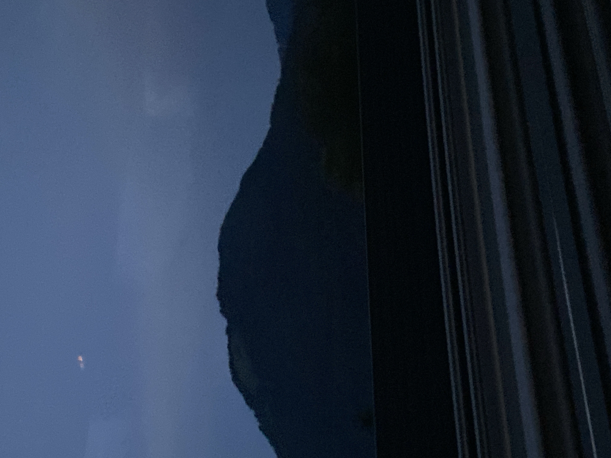 The image depicts a dark, mountainous landscape at dusk. The sky is a deep blue, with a hint of lighter shades near the horizon. In the upper portion of the sky, there are two faint, orange-hued lights, possibly distant aircraft or celestial bodies. A large, dark mountain range dominates the middle ground, silhouetted against the sky. The terrain appears to be covered in dense vegetation, though the details are obscured by the low light. In the foreground, there is a dark, horizontal structure, possibly a barrier or a bridge railing, with multiple horizontal lines suggesting a road surface or a textured material. The overall scene conveys a sense of twilight and tranquility. The location is identified as the city of Wörgl, Austria.