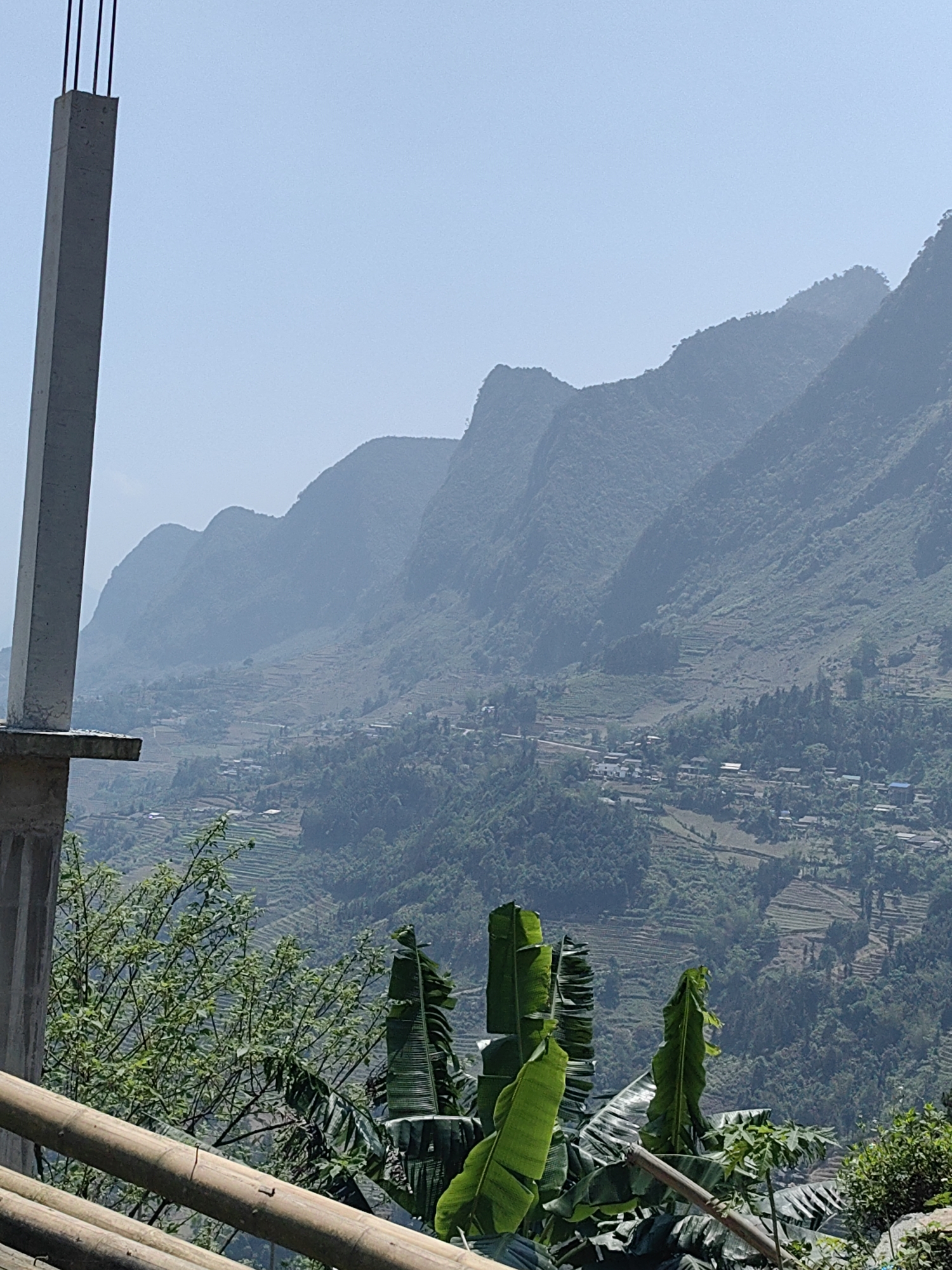 The image depicts a mountainous landscape in Yên Minh Commune, Vietnam. In the foreground, the corner of an unfinished concrete structure is visible. Green vegetation, including large banana leaves, grows near a bamboo railing. The midground consists of terraced fields and scattered houses along the slopes of the mountains. The background shows a series of mountain ranges under a hazy sky.