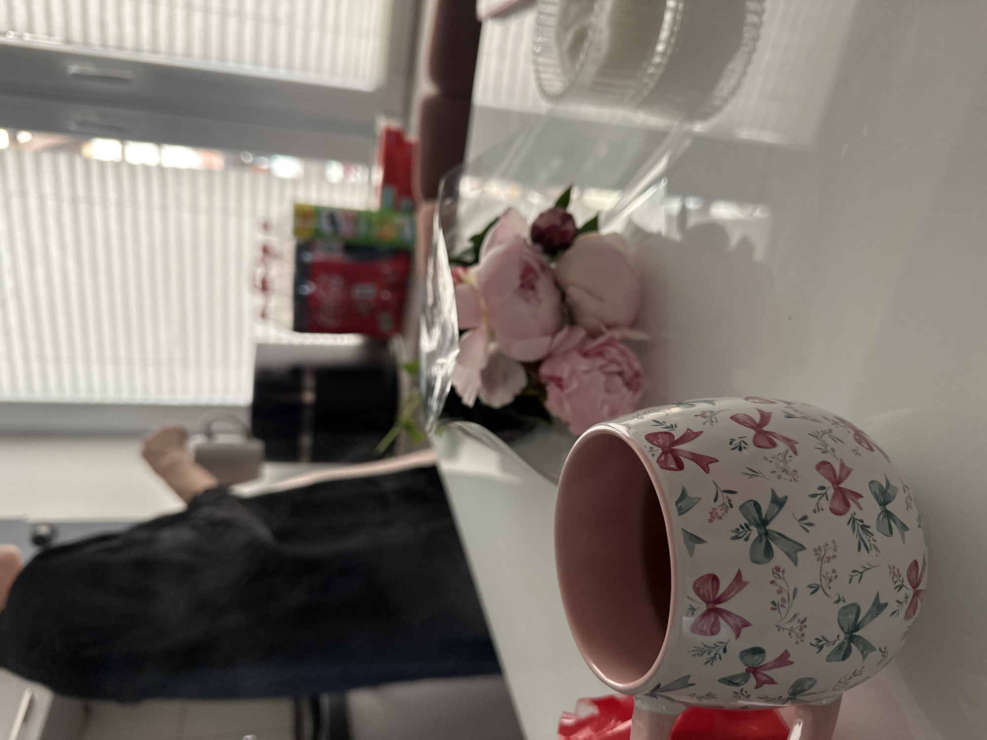 The image shows a close-up of a white table with a pink and white patterned mug filled with dark liquid in the foreground. The mug has a floral and bow design. To the right of the mug is a bouquet of pink peonies wrapped in clear plastic. In the background, a person in a black shirt is standing near a kitchen counter, holding a mug. There are bottles of Coca-Cola and Pringles on a shelf behind the person. The scene appears to be indoors, likely in a home kitchen or dining area, with blinds covering a window in the background. The lighting suggests it is daytime. There is no discernible information to link this scene to Kutenholz, Germany.