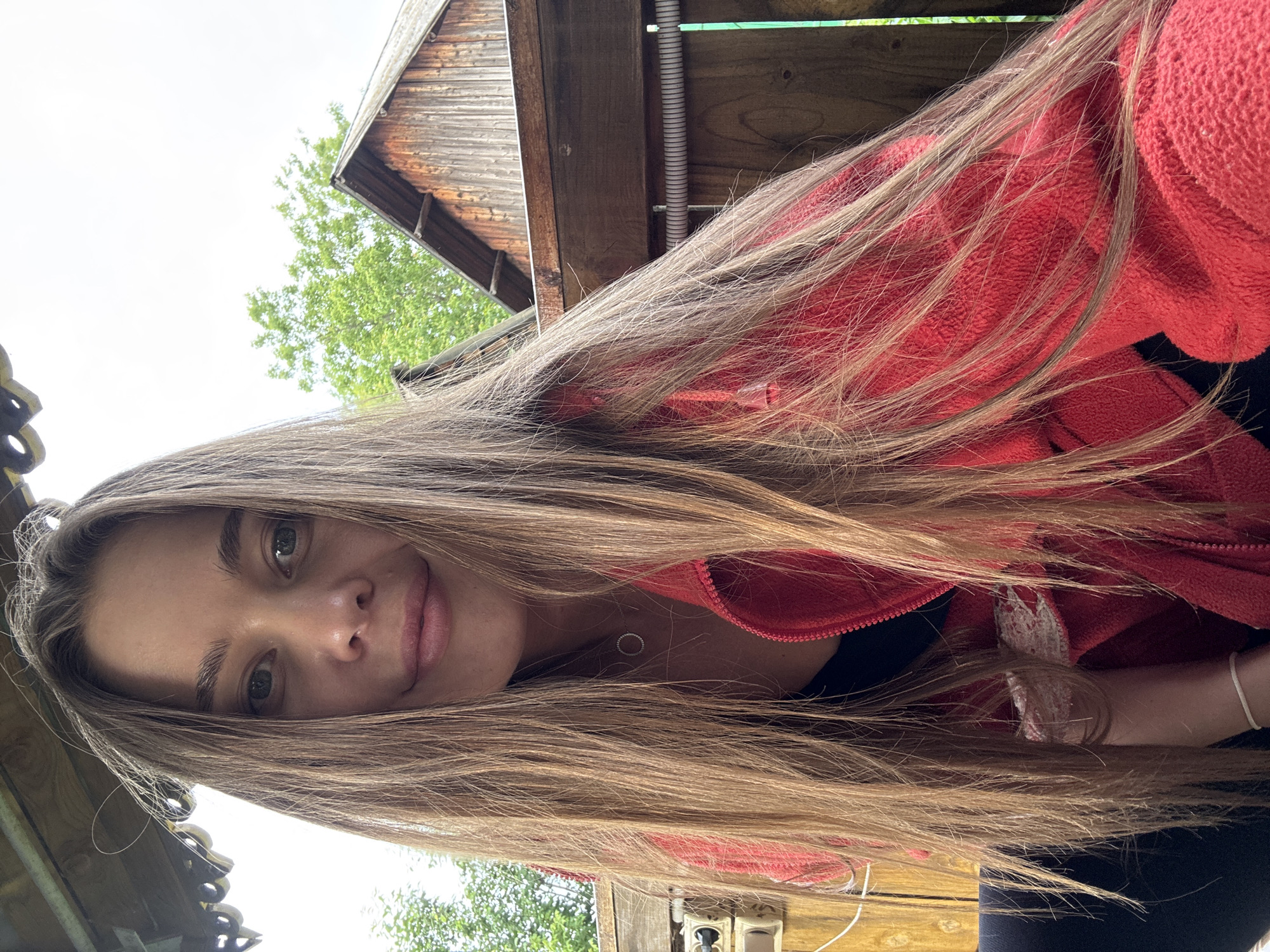 A young woman with long, blonde hair is taking a selfie outdoors. She is wearing a red fleece jacket over a black top and a delicate necklace. Her hair falls around her shoulders and obscures part of her face and body. The background suggests a rural setting, possibly in or near the village of Sulmash, Russia, with wooden structures and greenery visible. The overall lighting is bright, indicating daylight.
