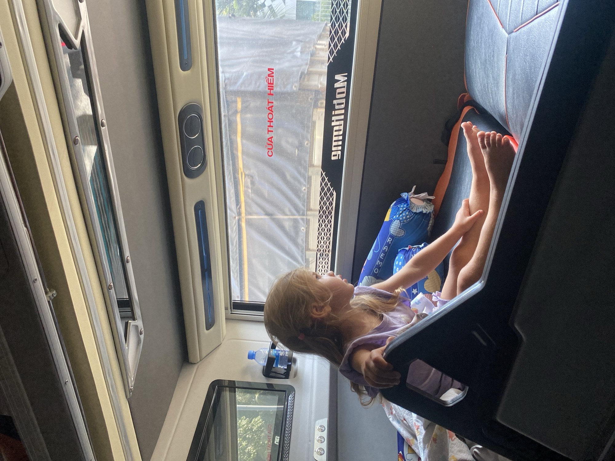 A young girl with blonde hair is sitting on a bed inside a vehicle, possibly a bus or van, looking out a window. She is wearing a purple shirt. The interior of the vehicle is visible, including the ceiling with vents and lights, and the window which shows a truck outside. There is a bottle of water on a shelf to the left of the girl, and a pillow with blue and white patterns on the bed. The text "CỬA THOÁT HIỂM" which translates to "EMERGENCY EXIT" is visible on the window. The overall environment suggests an indoor setting within a mode of transportation, likely in Ho Chi Minh City, Vietnam, given the Vietnamese text on the window.