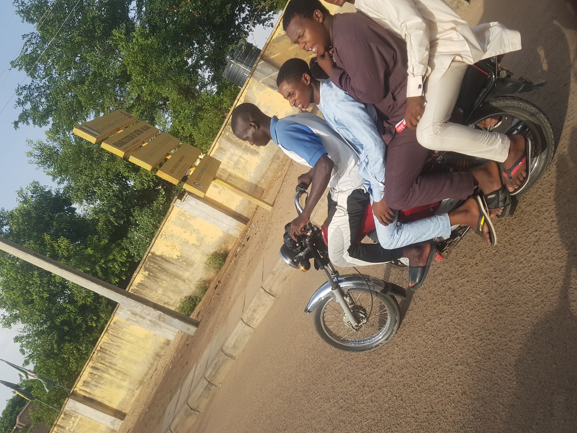 The image shows four young men riding on a single motorcycle on a paved road in Dutsinma A, Nigeria. The rider wears a blue and white shirt, while the three passengers wear a brown robe, a light blue shirt and light coloured pants respectively. None wear helmets or shoes. They appear to be travelling in front of a faded yellow building with a street sign attached to it. The environment is sunny and there are trees visible in the background.