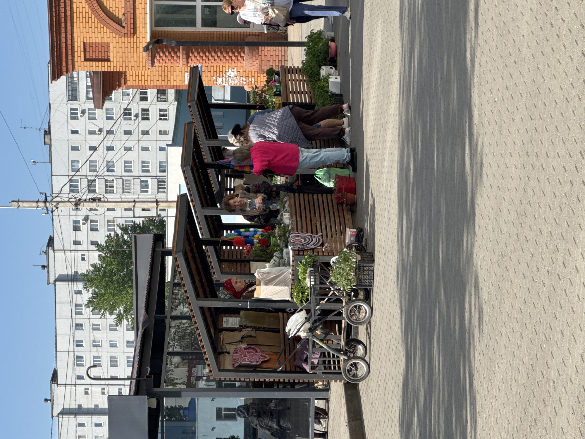 The image depicts an outdoor market scene in Chaikovsky, Russia. Several open-air stalls with dark wooden frames and roofs line a paved walkway. People are browsing and interacting with the vendors, examining items on display. One vendor is wearing a red headscarf. A stroller and a cart filled with goods are present near one of the stalls. In the background are multi-story residential buildings, a brick building, and utility poles. The time of day appears to be late morning or early afternoon, based on the shadows. The weather is clear and sunny.