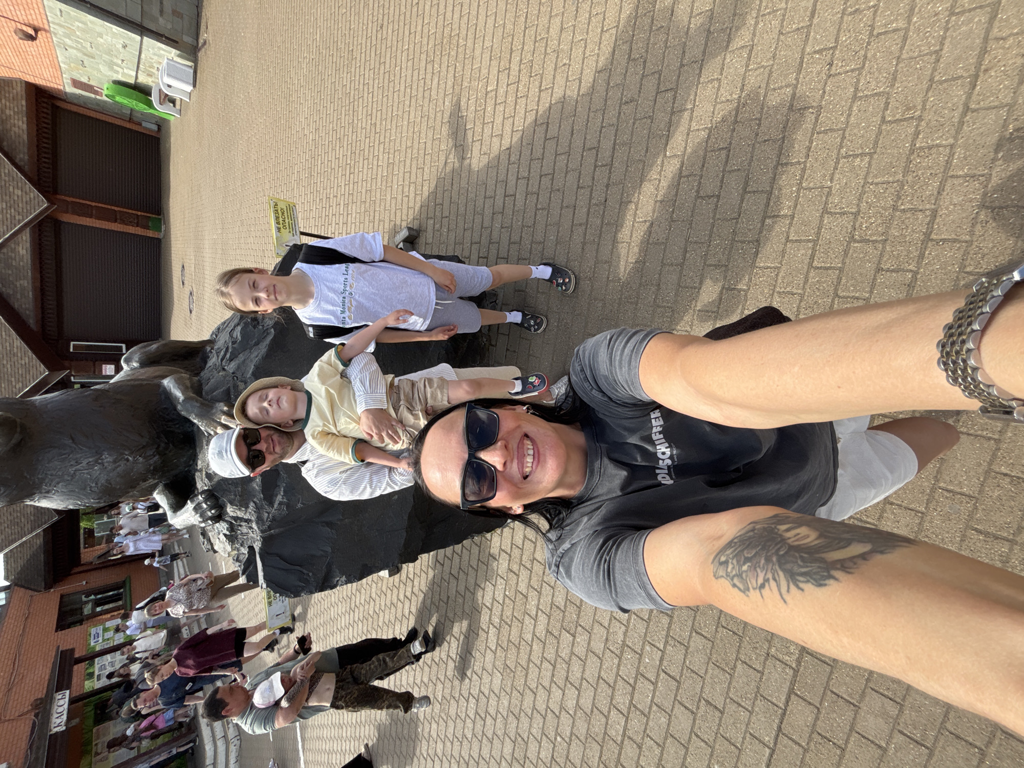 A selfie-style photograph, rotated counter-clockwise, shows a group of people posing near a monument. The foreground features a woman wearing sunglasses and a t-shirt with the word "DISCHIFER" printed on it, with a large tattoo on her left arm. Beside her are two men and two children. One man is wearing sunglasses and a white hat, and another is wearing a hat. One child has on a light-colored shirt and shorts, and the other child is wearing a light-colored hat.
In the background, numerous people are standing near buildings with storefronts. Signage with Russian lettering is visible on the buildings. The ground is paved with brick. The scene is well-lit, indicating daytime. Based on location, the setting is likely near the monument to Izhevsk Factory Workers in Izhevsk, Russia.