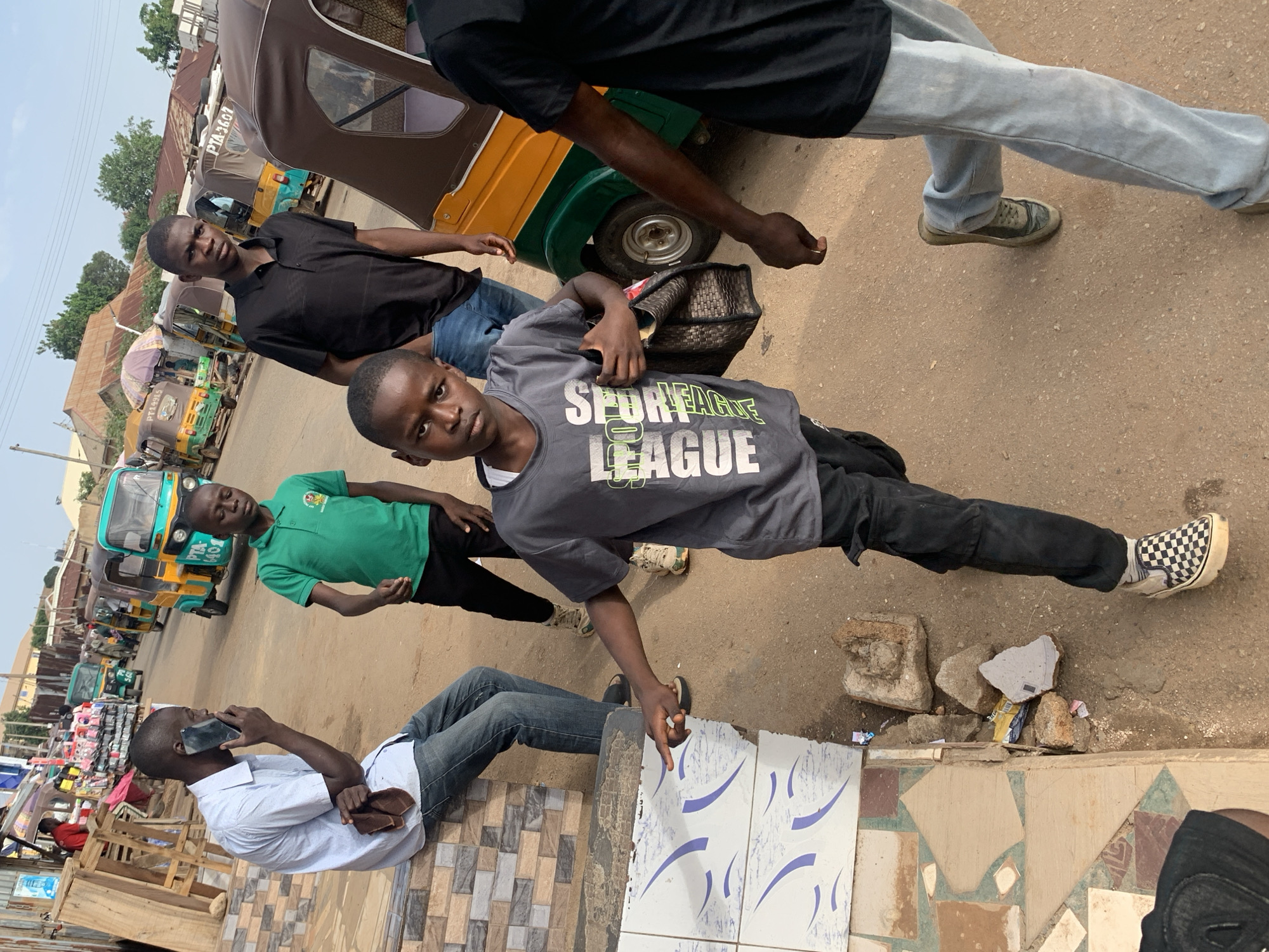 In Gura Top, Nigeria, a street scene unfolds with several individuals and vehicles. A man in a light blue collared shirt and jeans is seated on a low wall, holding a dark-colored mobile phone to his ear. Beside him, a young boy in a green polo shirt stands. In the foreground, a young boy in a gray t-shirt with white and green lettering, black pants, and checkered slip-on shoes is actively running, carrying a dark woven bag. Behind him, a man in a black collared shirt and blue jeans walks away from the camera. Further in the background, several auto-rickshaws, characteristic of Nigerian transportation, are parked and in motion. The environment appears to be a dusty street with some market stalls and buildings visible in the distance. The overall atmosphere suggests a typical day in a bustling urban area.