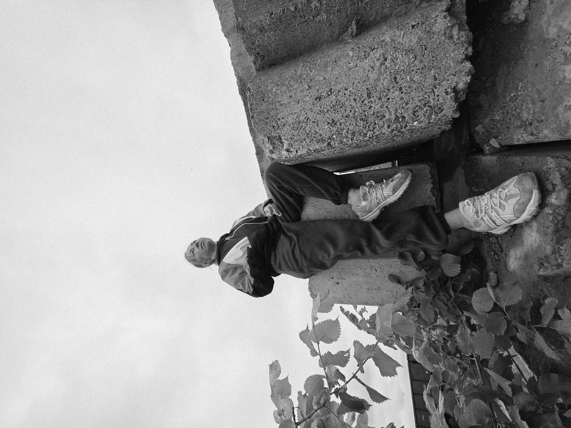 The image, taken in Chaikovsky, Russia, features an adult individual, likely female, seated on a large, roughly textured concrete block. The person wears a tracksuit, consisting of a dark and light-paneled zip-up jacket over a dark layer, dark tracksuit bottoms, and light-colored athletic shoes. Her head is tilted upwards, and she gazes towards the upper right of the frame. Her left foot rests on a lower concrete surface, while her right foot is positioned on the block she is seated on. The upper portion of the scene is dominated by a bright, overcast sky. A prominent leafy branch extends from the lower left foreground into the mid-ground. To the right and below the seated individual, additional large, unhewn concrete blocks with visible aggregate are present. In the far left background, partially obscured by foliage, a dark, horizontal structure, consistent with a train car or industrial building, is visible. The immediate ground around the base of the concrete blocks appears uneven and unpaved.