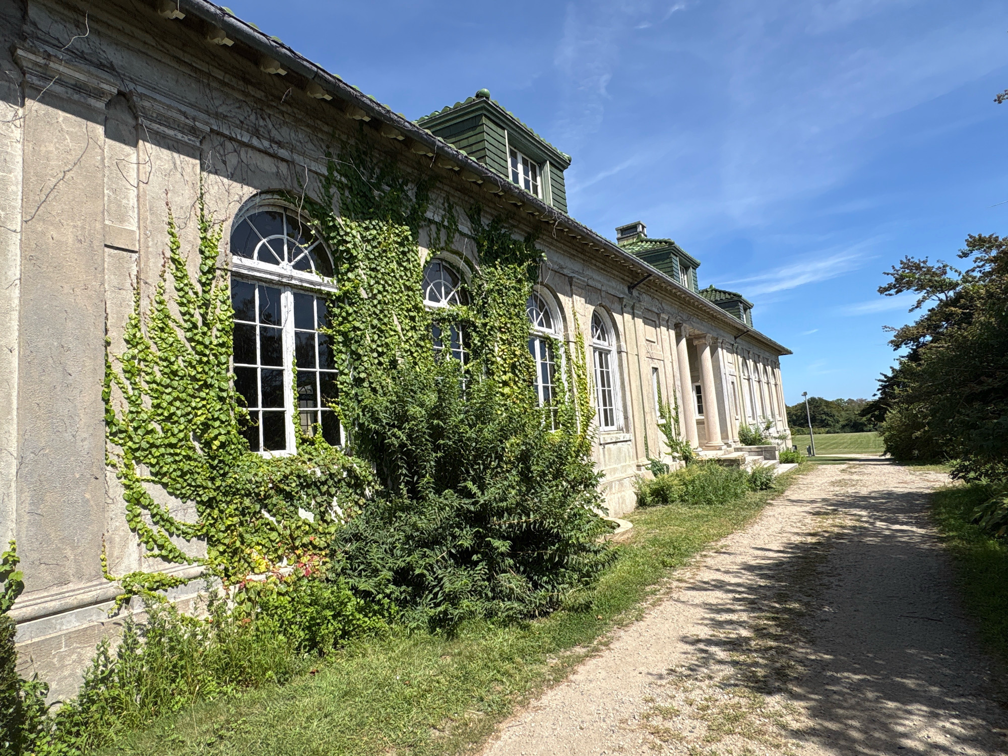 The image captures the side of a stone building with green ivy climbing its walls and covering some of its windows. The building features several arched windows with white frames. The roof is covered in green tiles and has dormer windows. A dirt path runs alongside the building, bordered by grass and trees. In the background, a green field extends towards the horizon under a blue sky with scattered white clouds. The scene is devoid of any people. This location is identified as Waterford, United States of America.