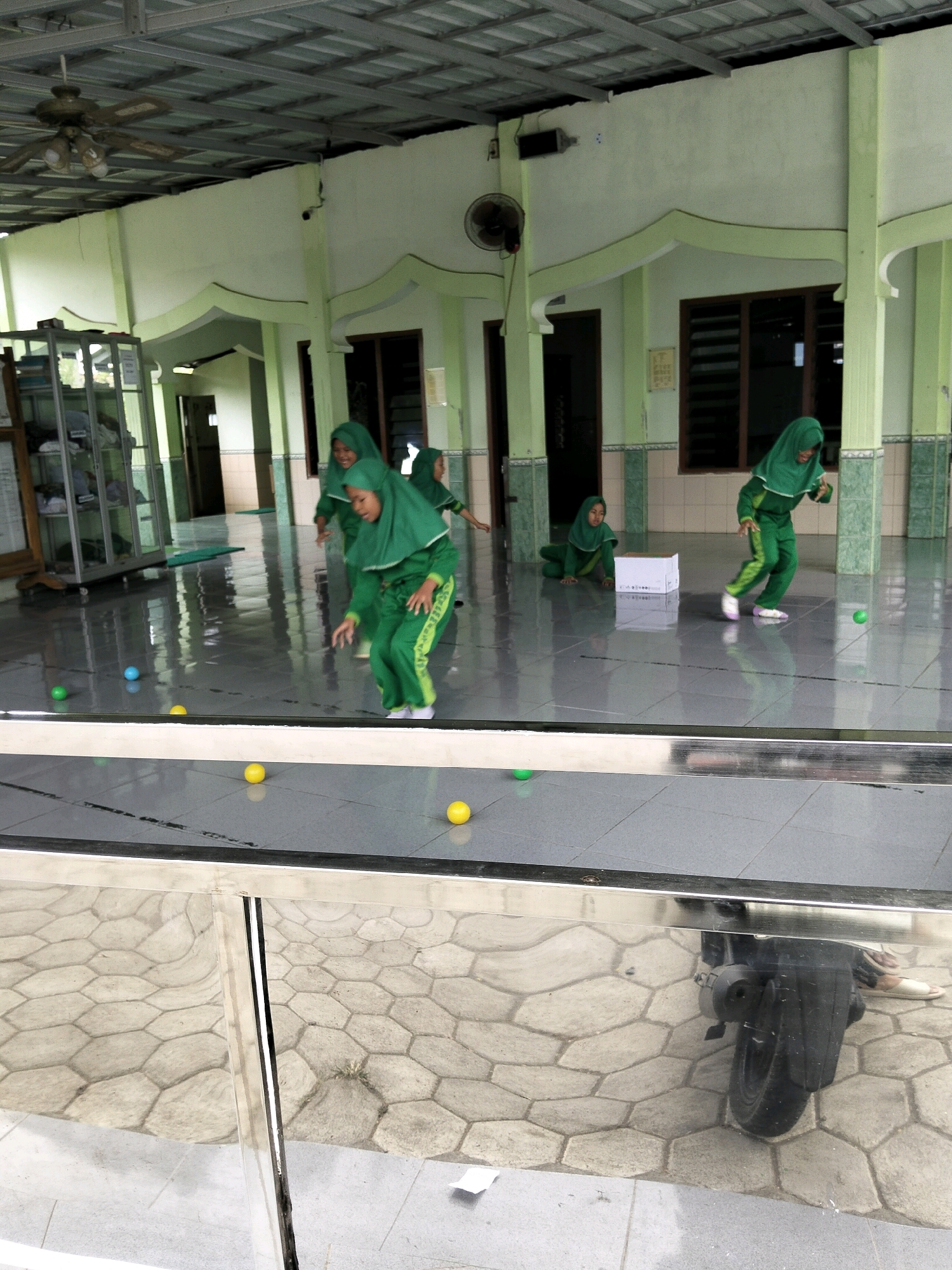 In Kemiri Kidul, Indonesia, within an open-air structure with a metal roof supported by light green pillars, five young girls wearing green hijabs and uniforms are engaged in an activity on a polished concrete floor. The girls are spread across the space, with some in the foreground and others further back. One girl on the far right is in motion, appearing to throw a small green ball. To her left, another girl is on her knees beside a white box. Further back and to the left, three more girls are positioned, one of whom is also reaching out. Scattered on the floor are several small, colorful balls, including green, yellow, and blue. In the background, a glass cabinet filled with items is visible, as are large windows and doorways leading to other areas. A ceiling fan is mounted overhead. A reflective metallic railing runs across the foreground, and the reflection of a motorcycle wheel is visible on its surface.