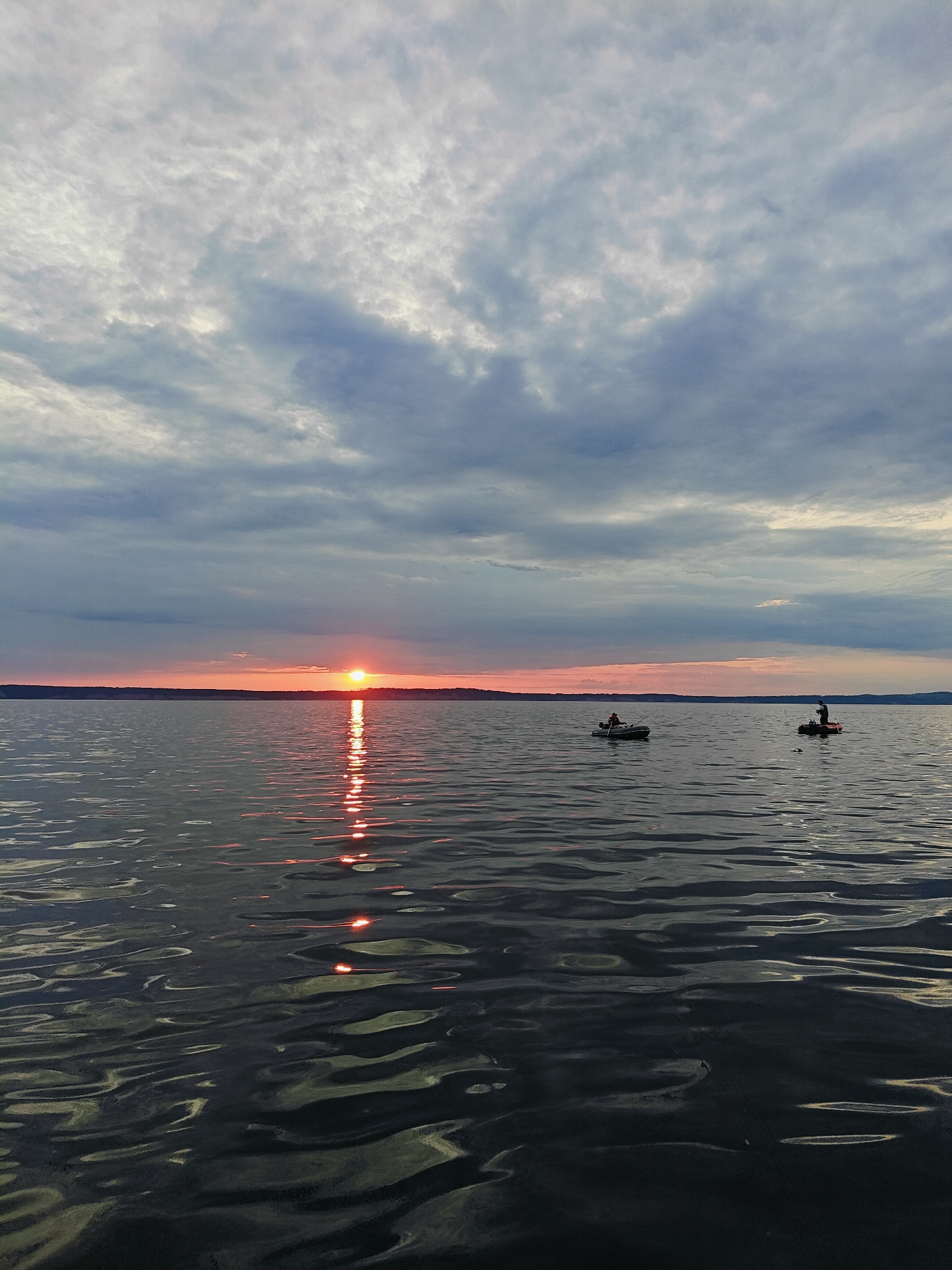 The image captures a sunset over the Kama River in Chaykovsky, Russia. The sky is filled with a mix of light and dark clouds, with the setting sun casting a warm orange glow near the horizon. The sun's reflection creates a shimmering path of light across the calm water. In the distance, a thin strip of land, likely the shore, is visible. Two small boats are present on the water. One boat, closer to the left, has a single figure who appears to be fishing. Another boat, further to the right, also contains a single person, who seems to be standing and possibly fishing as well. The water exhibits gentle ripples, reflecting the muted colors of the sky and the vibrant sunset.