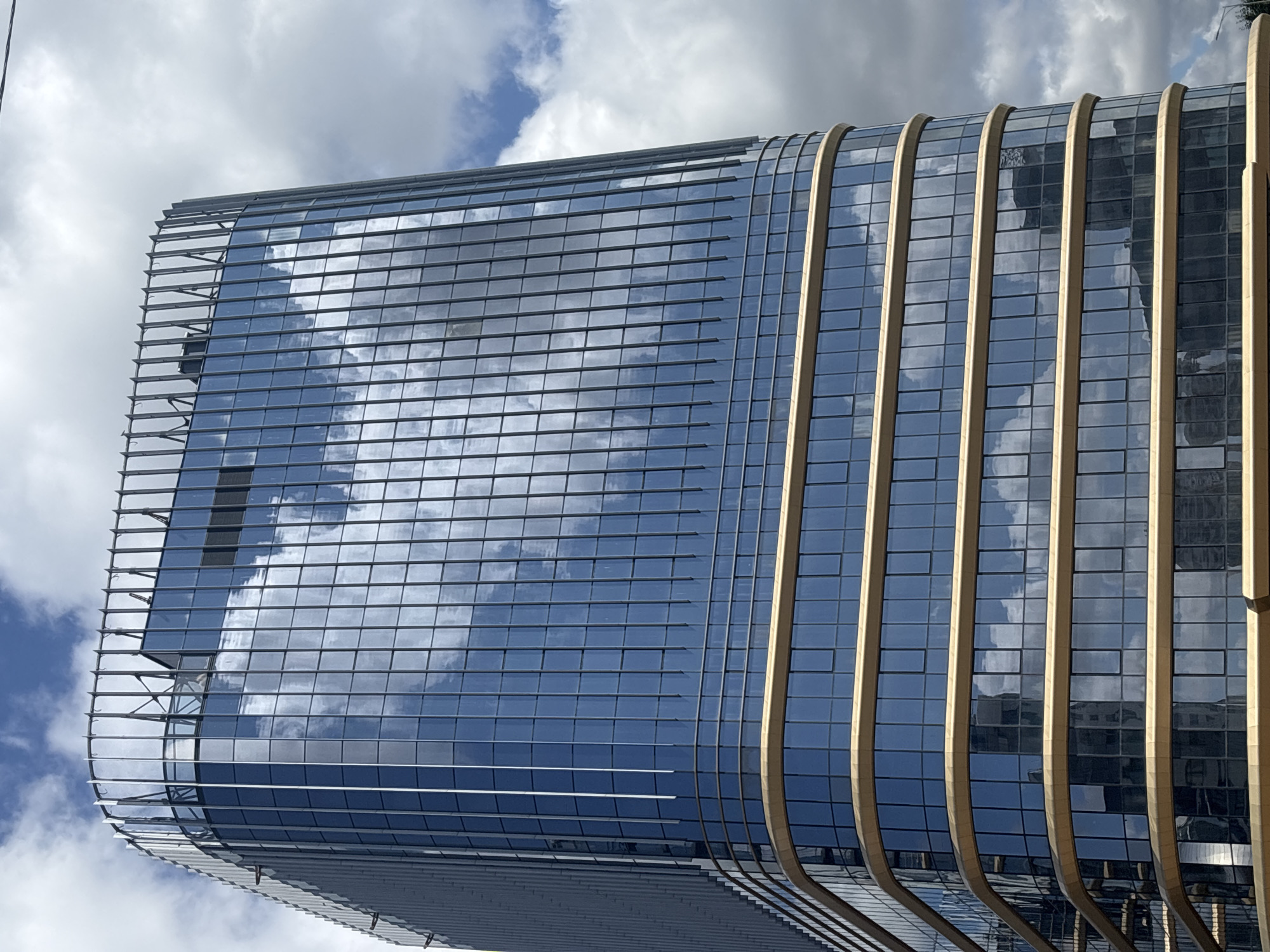 A modern glass skyscraper with a curved facade and gold accents reflects the sky and clouds. The building features a grid of rectangular windows and horizontal gold banding at different levels. A light gray metal railing structure crowns the top of the building. The sky above is partly cloudy with white and gray clouds against a blue sky. The image appears to be taken from a low angle, looking up at the building. The building is located in Minsk, Belarus.