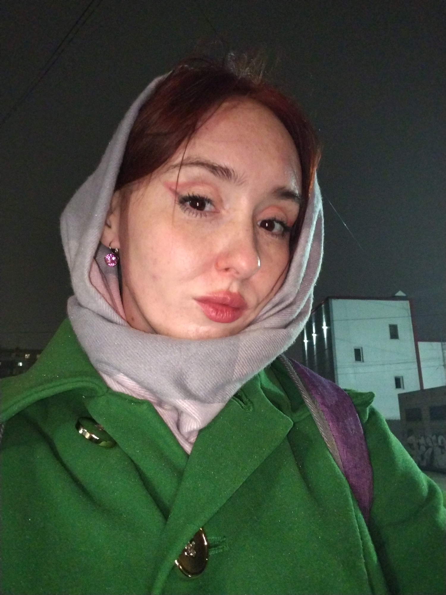 A female presents a selfie in an outdoor setting, presumably at night in Izhevsk, Russia, judging from the architecture in the background. She wears a green coat with gold-colored fasteners, a pink scarf, and a light grey head covering. A purple backpack strap is visible over her shoulder. The woman has red hair and pink eyeliner. She is wearing a pink earring in her left ear. A building is visible in the background.