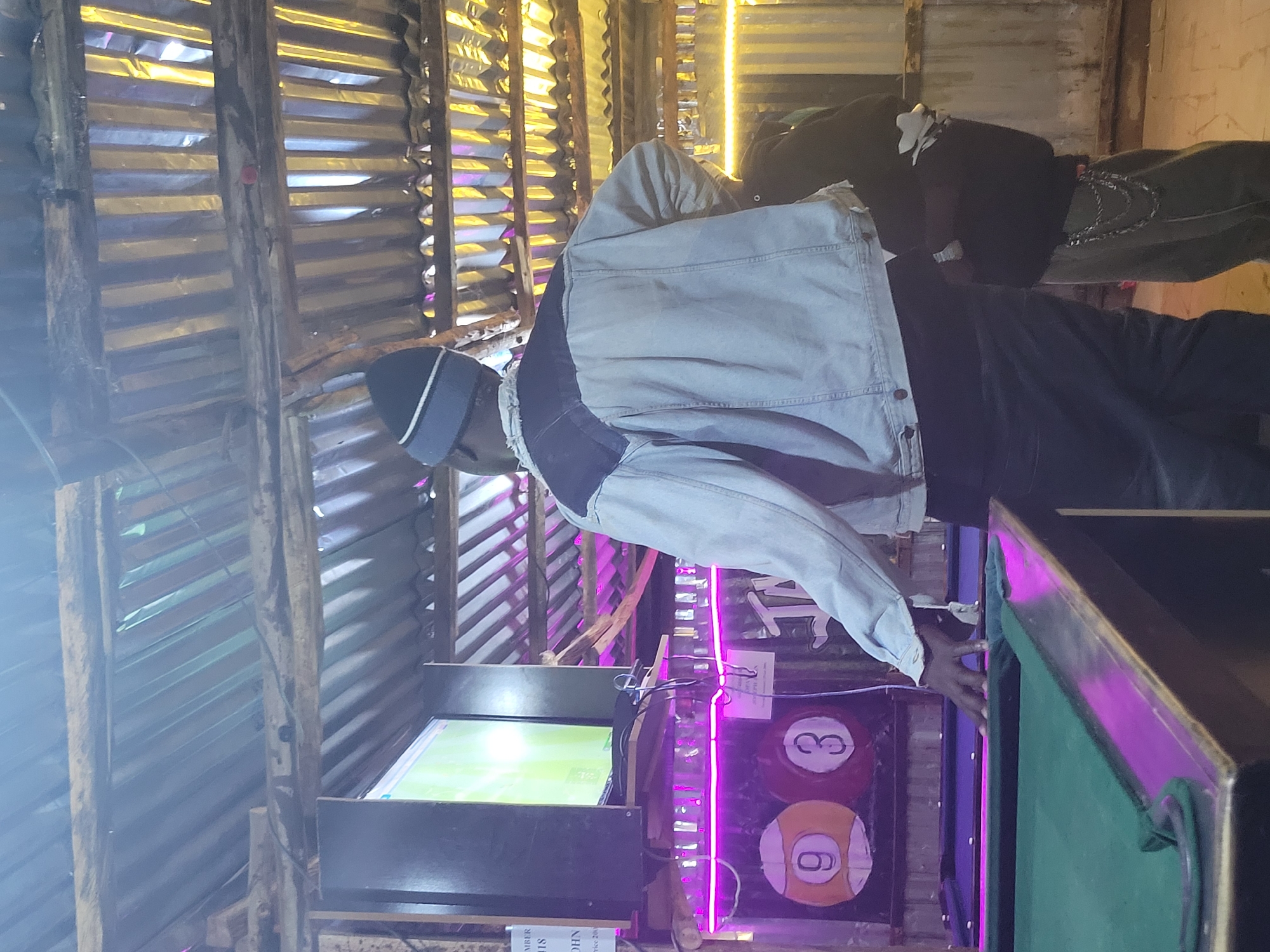 In Durumi, Nigeria, a man wearing a denim jacket and a beanie stands with his back to the camera, facing a pool table. His right hand is on the edge of the green felt table. To the left of the pool table, a television screen displays a bright green image, and two pool balls, numbered 9 and 3, are visible painted on the wall behind the table. The environment appears to be an indoor space with a corrugated metal roof and neon purple lighting. Another person, partially visible, stands to the right of the main subject. The overall scene depicts a casual setting, likely a bar or recreation center.