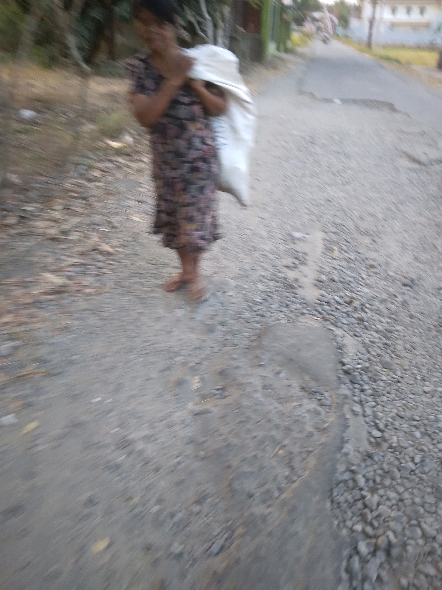 The image depicts a person carrying a white sack over their shoulder while walking on a gravel road in Desa Pamijen, Kecamatan Sokaraja, Indonesia. The person is wearing a dark floral dress and appears to be barefoot. The road is uneven and covered in small stones and dirt. To the left of the road, there is dry vegetation and some trees. In the background, a paved road is visible with some buildings and vegetation. The lighting suggests it is daytime, possibly late afternoon due to the soft light and shadows. The overall scene is rural and appears to be a residential or agricultural area.
