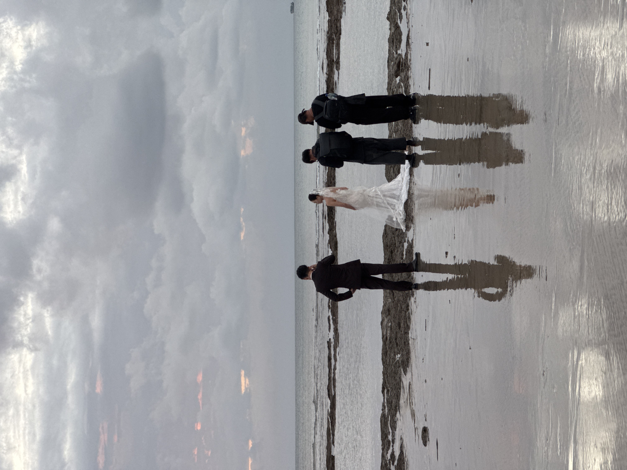 A wedding ceremony is taking place on a beach in Kuta Selatan, Indonesia. Four people are present: a bride in a white wedding dress and veil, and three men in dark suits. The bride is facing away from the camera, bowing her head. Two of the men are also bowing, facing the bride. The third man is standing to the left, facing away from the camera, with his hand on his head. The sky is cloudy and overcast, suggesting it is either early morning or late afternoon. The sun is partially visible, casting a faint glow through the clouds. The beach is wet, reflecting the figures of the people and the sky. There are patches of sand and rocks visible along the shoreline. The overall mood appears solemn and reflective. No text is visible in the image.