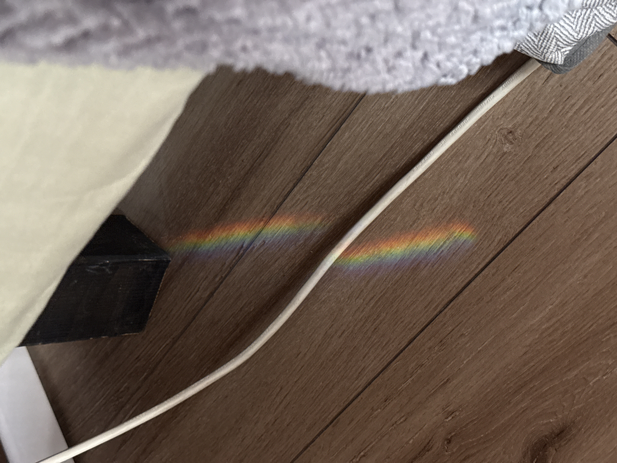 This image depicts an indoor scene in the city of Tel-Aviv, Israel, featuring a wooden laminate floor, a white electrical cable, and two distinct prismatic light reflections. The floor consists of medium-brown planks laid diagonally across the frame, exhibiting a visible wood grain pattern. A white, cylindrical cable runs from the bottom left, ascending towards the upper right, bearing the legible inscription: "SOMEBODY YKUXH IEC 2020-03 E02292-E".
Two vertical bands of spectral colors, resembling rainbows, are cast upon the floor. The upper rainbow is partially obscured by a dark, rectangular object, likely a furniture leg, positioned beneath a light beige fabric surface, possibly a bed skirt or sofa cover, visible in the upper right quadrant. The lower rainbow is located further down the floor, slightly to the left of the white cable. Along the right edge of the frame, a textured, light grey, possibly knitted or woven fabric, such as a blanket or rug, is visible, partially covering the white cable at the bottom right. Adjacent to this textured fabric, where it meets the cable, is a strip of fabric displaying a light and dark grey herringbone or chevron pattern. A white baseboard or wall section is visible in the upper left corner. No individuals or explicit actions are present in the frame.