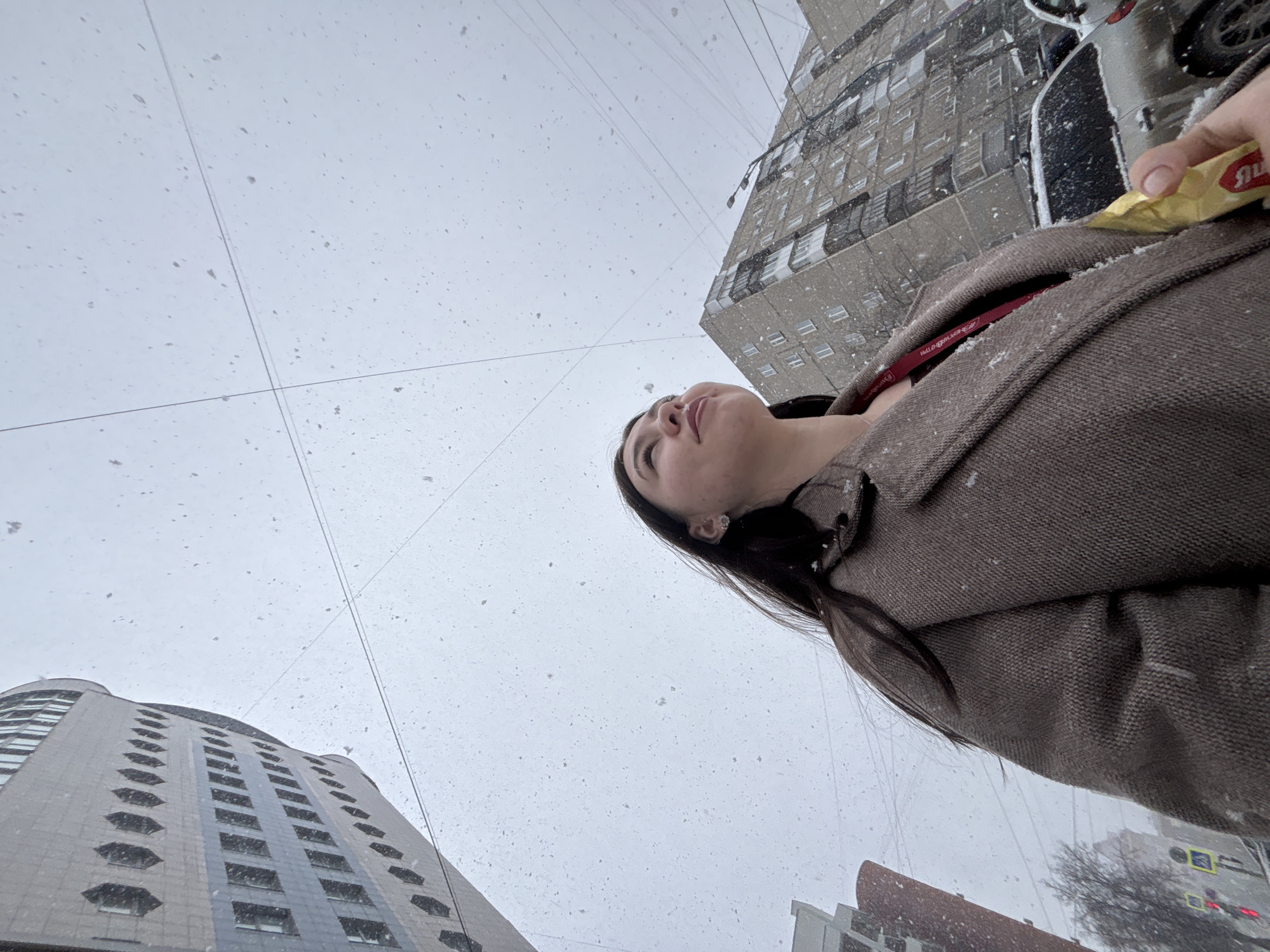 A woman stands on a snowy street in Perm, Russia, looking up at the sky. The camera captures her from a low angle. She wears a brown coat, and her dark hair is visible. The backdrop includes multiple buildings, and snowflakes are visible in the air. Her hand is near a yellow package, and part of a vehicle is visible on the right. Overhead lines run across the sky.