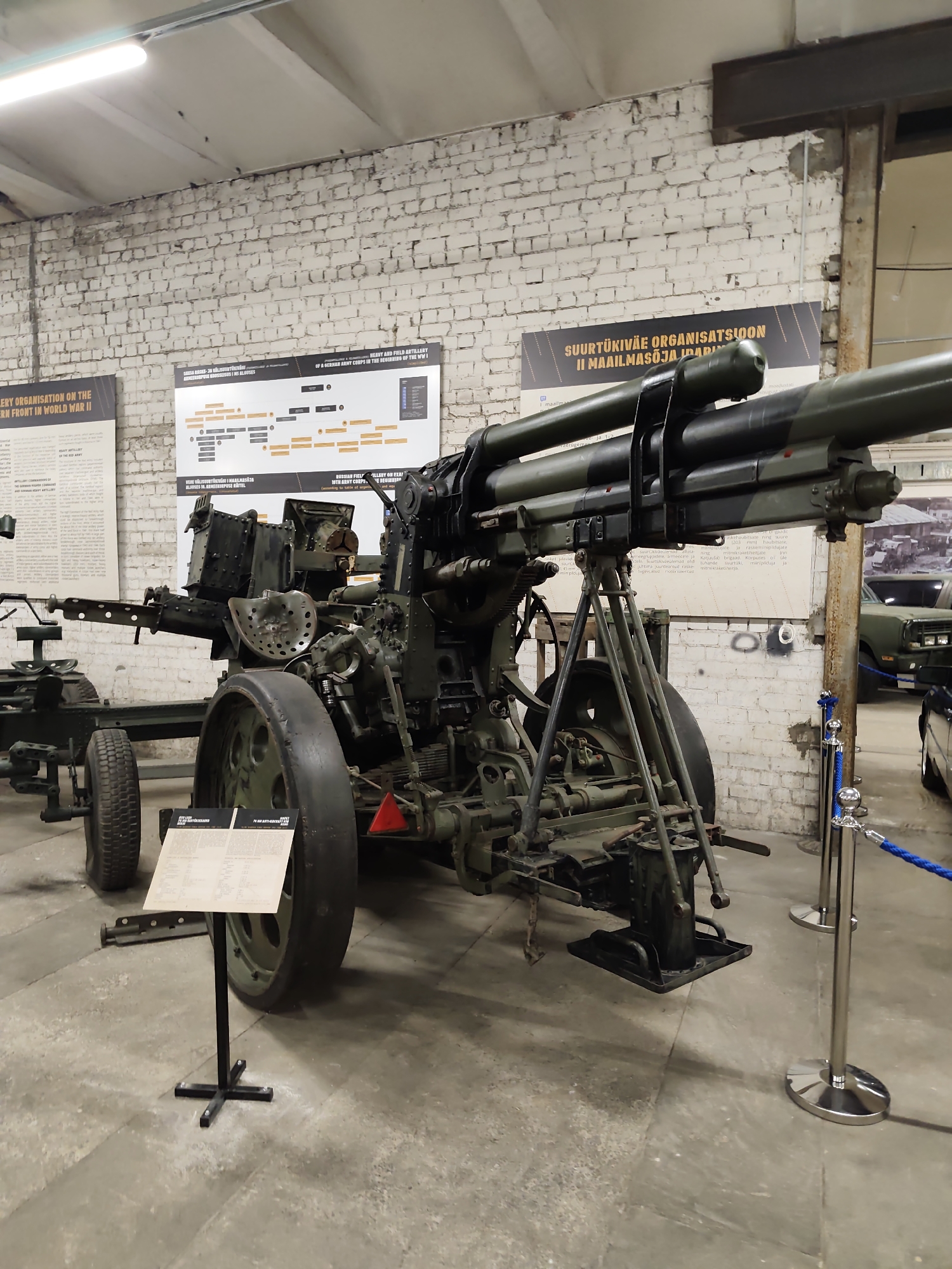 This media file is a photograph of a historical military exhibit, likely in a museum. The primary subject is a large, dark green artillery piece, possibly an anti-aircraft gun, displayed in what appears to be an indoor exhibition hall with brick walls and a concrete floor. The gun is complex, with multiple barrels, a wheeled carriage, and supporting legs.
To the left of the main artillery piece, another, less detailed military vehicle or gun carriage is partially visible. In the background, there are several informational posters displayed on the brick wall, providing historical context. Some text on these posters is partially legible. One poster has a title that appears to be in Finnish or a similar language, mentioning "SUURTÜKIVÄE ORGANISATSIOON II MAAILMASÕJA". Another poster is in English and discusses "ARTILLERY ORGANISATION ON THE WESTERN FRONT IN WORLD WAR II" and "HEAVY AND FIELD ARTILLERY OF A GERMAN ARMY CORPS IN THE DEGINNING OF THE WW I".
In the far background, parts of what look like military vehicles are visible, and a blue rope barrier guides visitors. The lighting suggests an indoor setting, likely during the day. The overall mood is educational and historical, focusing on military history. The location is identified as Viimsi, Estonia.