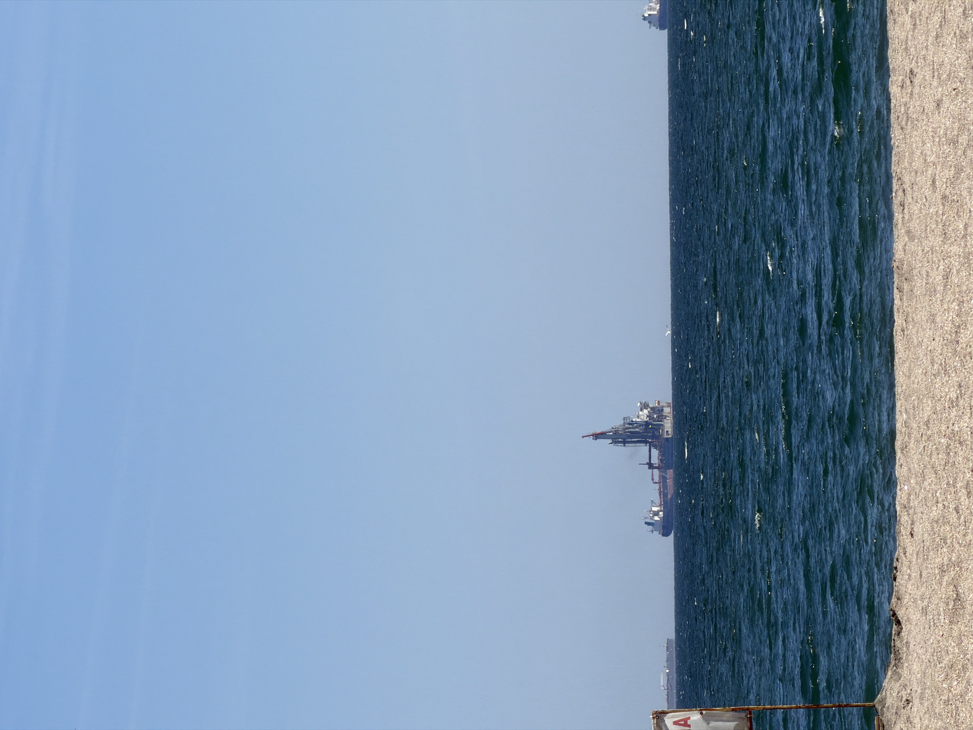 The image shows a seascape with a clear blue sky dominating the view. A body of water, possibly an ocean or a large sea, is visible with dark blue, slightly choppy waves. A light-colored, sandy or gravelly beach borders the water on the right. In the distance, several ships or industrial structures are visible along the horizon. At the bottom of the image, a portion of a weathered barrier or fence is present, with some markings or lettering visible on its surface. The location appears to be a coastal area, although the city is unknown.