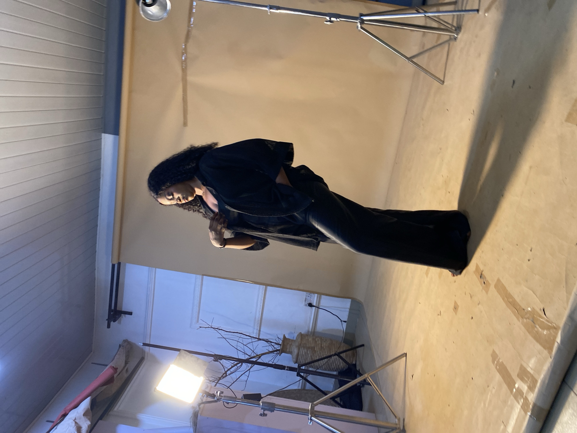A young Black woman with dark, curly hair is posing for a photo shoot in a studio setting. She is wearing a black jacket and black pants. The background consists of a large, light brown seamless paper backdrop. In the foreground, there are several pieces of studio equipment, including light stands and a portable photography light. A dried branch is positioned in front of a tall, woven vase. The lighting suggests an indoor setting, likely a studio. The overall mood appears to be professional and artistic. The time of day and weather are not discernible from the image. No visible text is present. The location context is Bukuru, Nigeria.