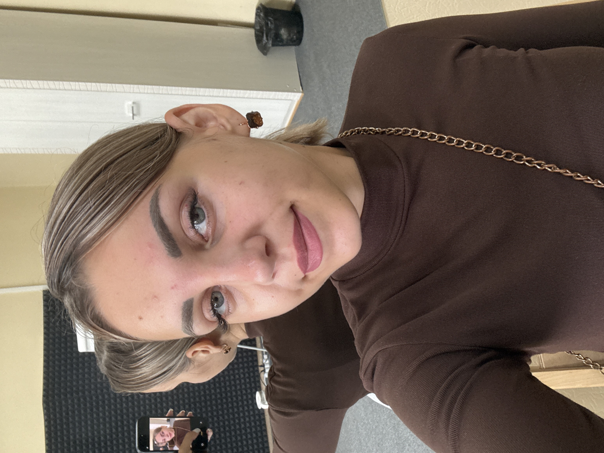 A young woman with light brown hair styled in a low ponytail with blonde highlights, wearing a brown turtleneck shirt and a gold chain necklace, takes a selfie in what appears to be a room with beige walls and a grey carpeted floor. She is positioned in the foreground, her face angled slightly to the left, with blue eyes and dark eyebrows. Her lips are painted with a matte pink lipstick. She is wearing small, brown, square-shaped earrings. In the background, a large mirror reflects her and the room. To the left of the mirror, a black acoustic foam panel is visible on the wall. To the right, a white door with a silver handle is ajar, revealing a sliver of the room beyond. A black trash can is situated near the wall to the right of the door. The woman is holding a smartphone in her left hand, which is capturing her reflection in the mirror. The overall lighting suggests an indoor setting, possibly a home or studio, on a neutral day. There are no obvious indicators of Yuzhno-Sakhalinsk, Russia.