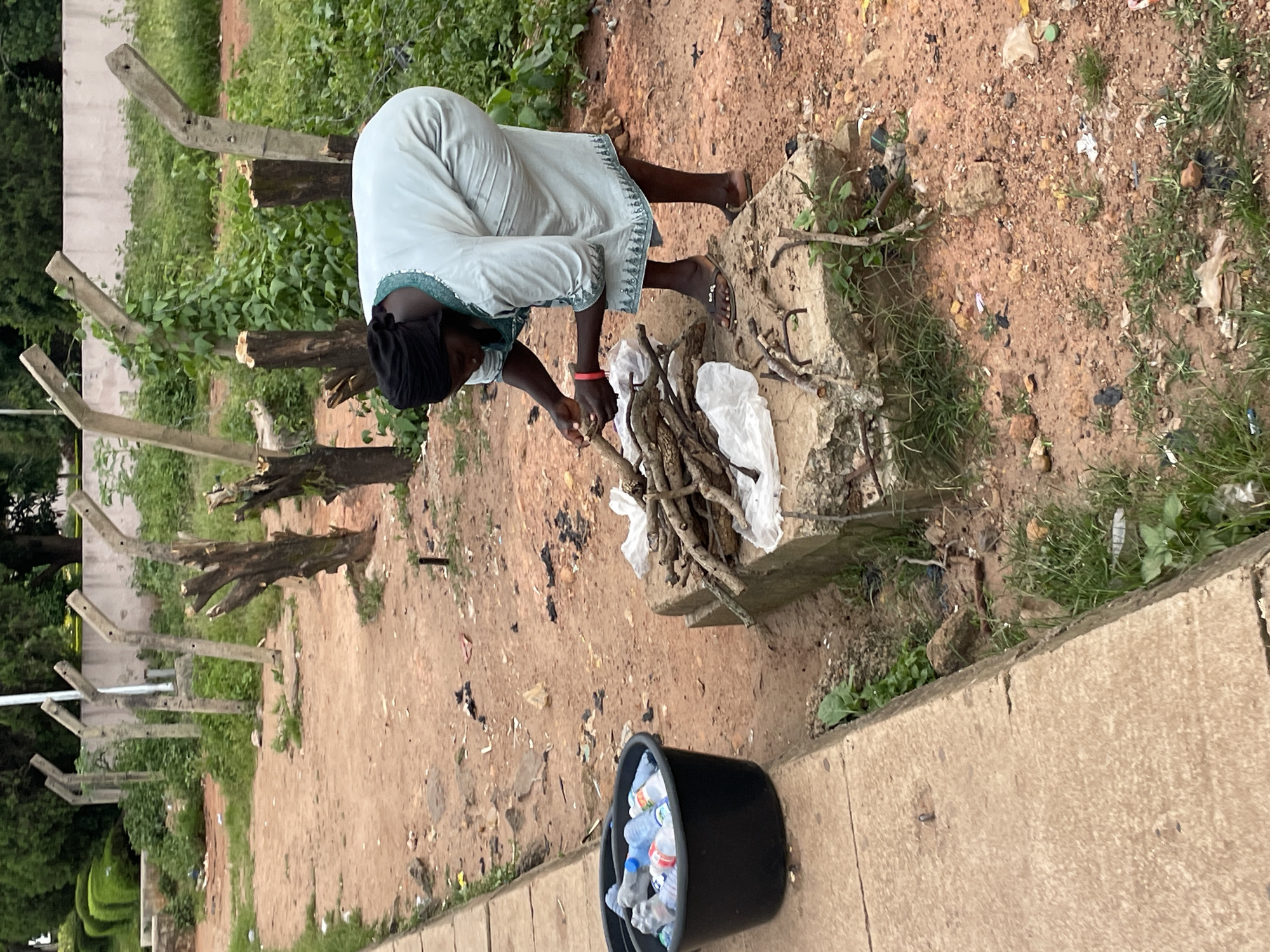 A woman wearing a light blue, long-sleeved dress and a black headscarf is in the process of arranging sticks on a concrete block in a dirt and grass area. She is bending over, holding several sticks in her hands. Beside her is a black bucket filled with plastic water bottles. In the background, there are several weathered concrete posts with angled tops, some with wire attached. The ground is covered in dirt, pebbles, and scattered debris, including plastic bags and remnants of burnt material. Lush green vegetation, including bushes and grass, grows around the posts and in patches on the ground. The overall scene appears to be an outdoor area within a city, possibly on the outskirts or in a less maintained part of Jos, Nigeria.