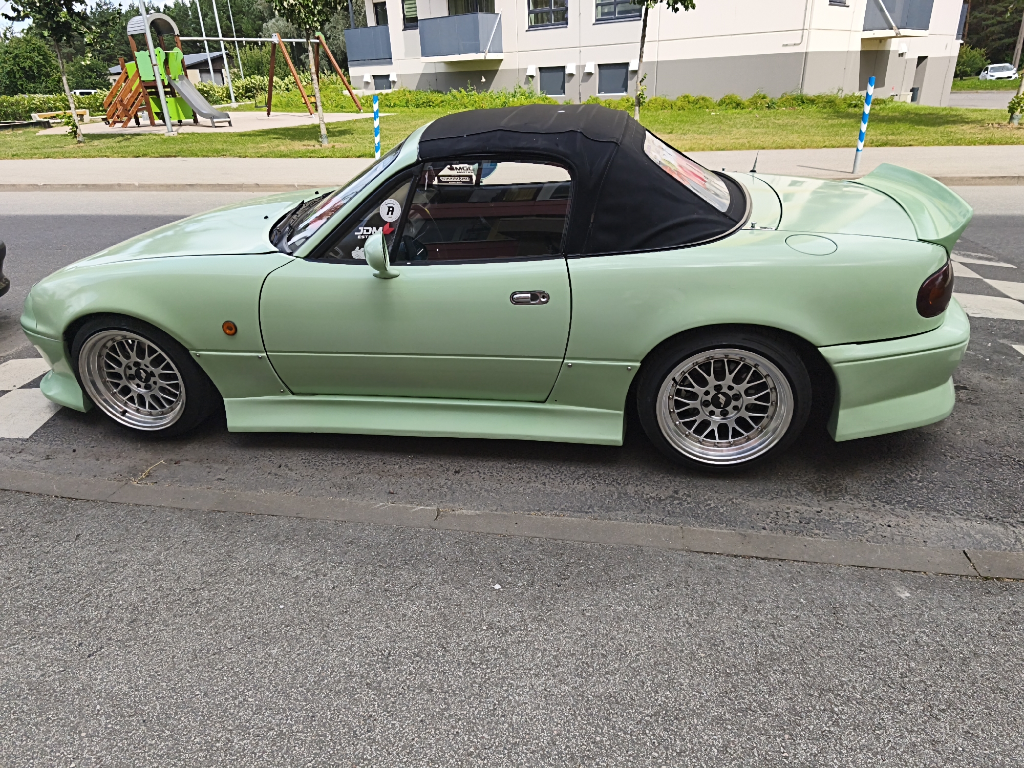 A light green convertible sports car, possibly a Mazda Miata, is parked on a street in Tallinn, Estonia. The car has a black soft top and aftermarket wheels. It is positioned on an asphalt road with a grey concrete curb. In the background, there is a playground with wooden and green structures, a sidewalk, and a multi-story building with grey and white facades. Trees and green foliage are also visible. The weather appears to be overcast.